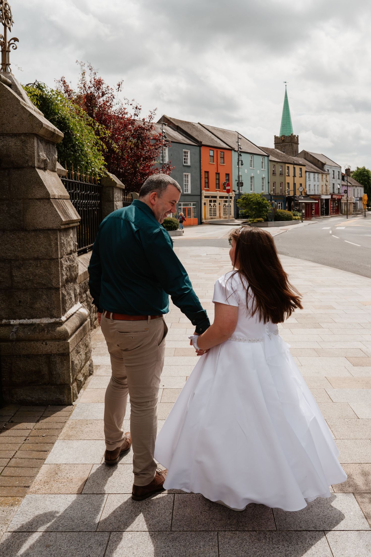 Man in green shirt holds hands with girl in white communion dress walking along colorful Irish town street.