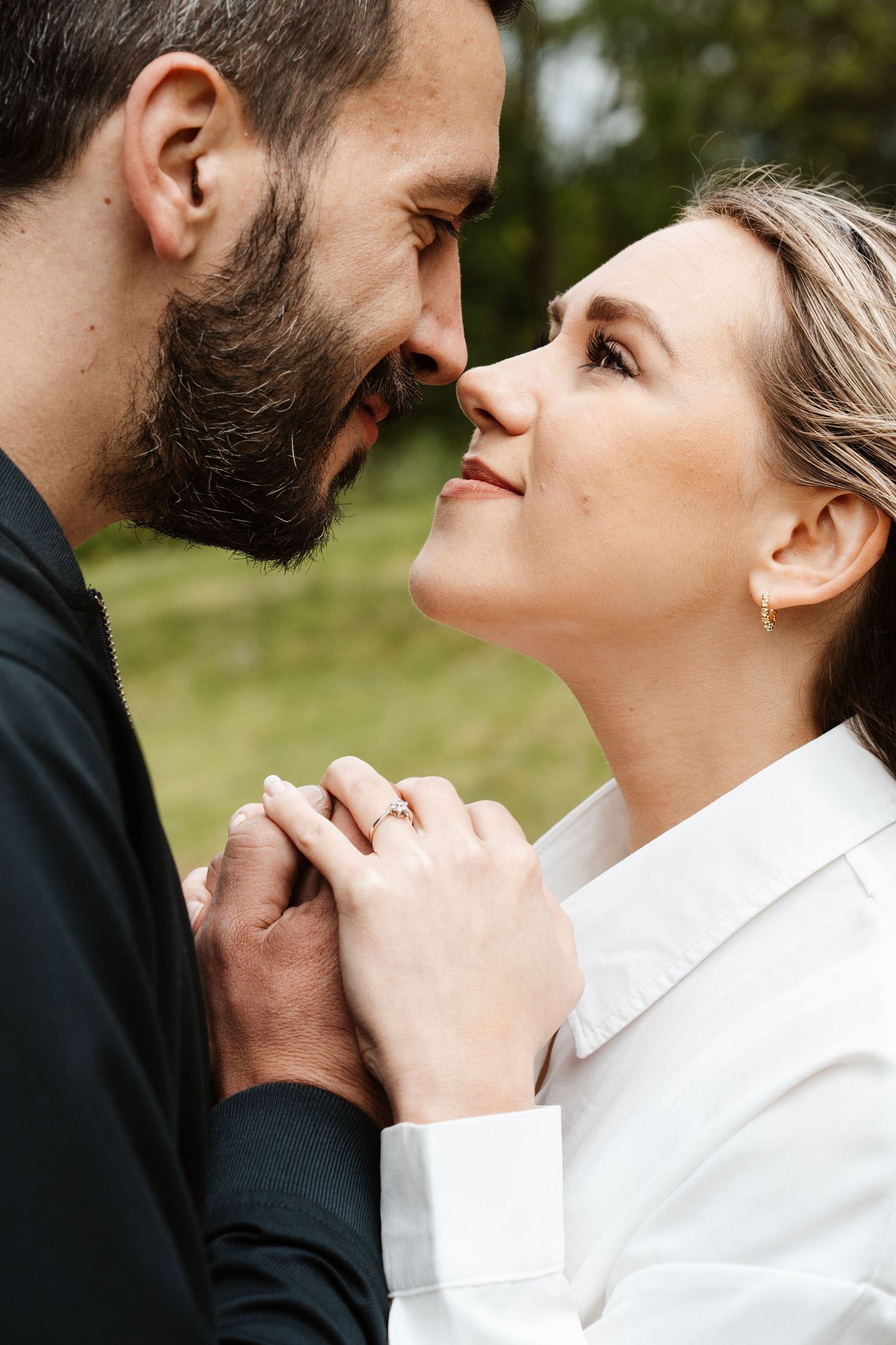 Romantic couple nearly kissing outdoors, woman wearing engagement ring, holding hands tenderly in a green park setting.