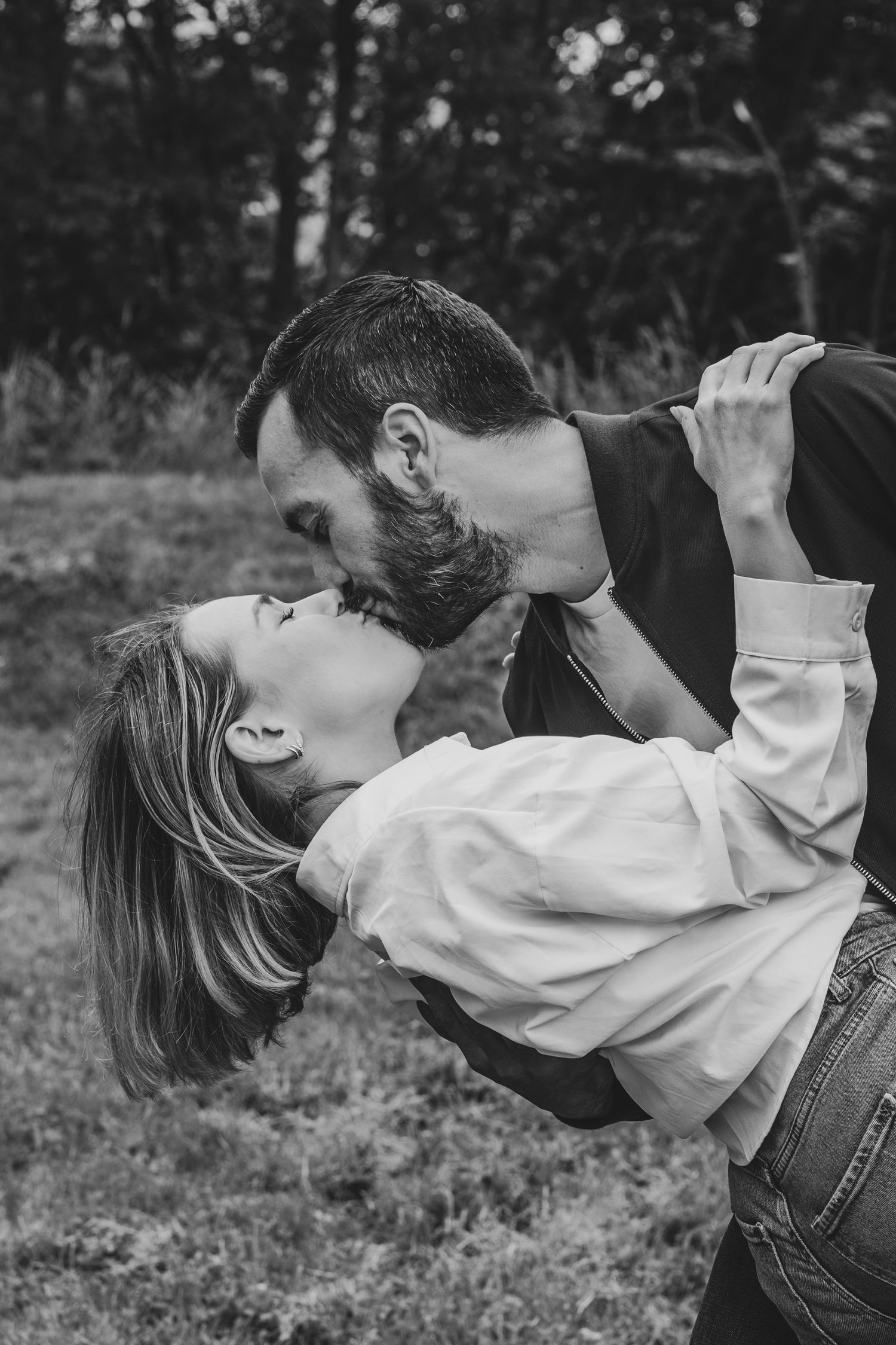 Black and white photo of a couple sharing a romantic dip kiss outdoors with trees in the background.
