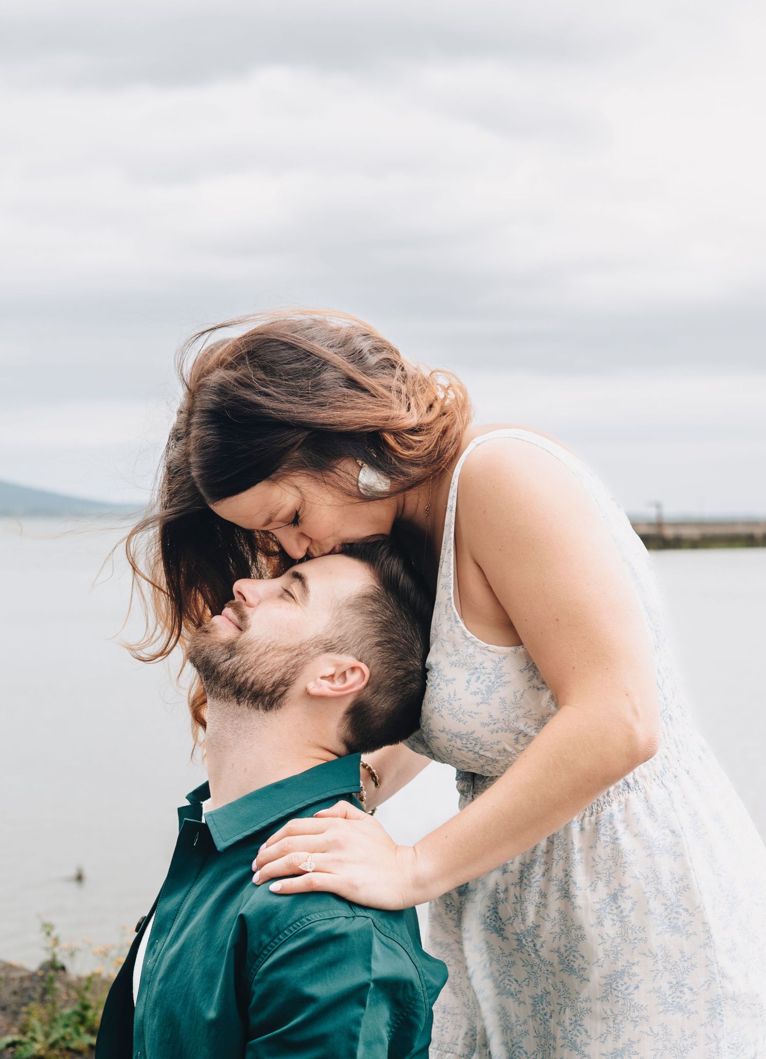 Romantic couple sharing a tender kiss outdoors near a lake on an overcast day, woman in a floral dress.