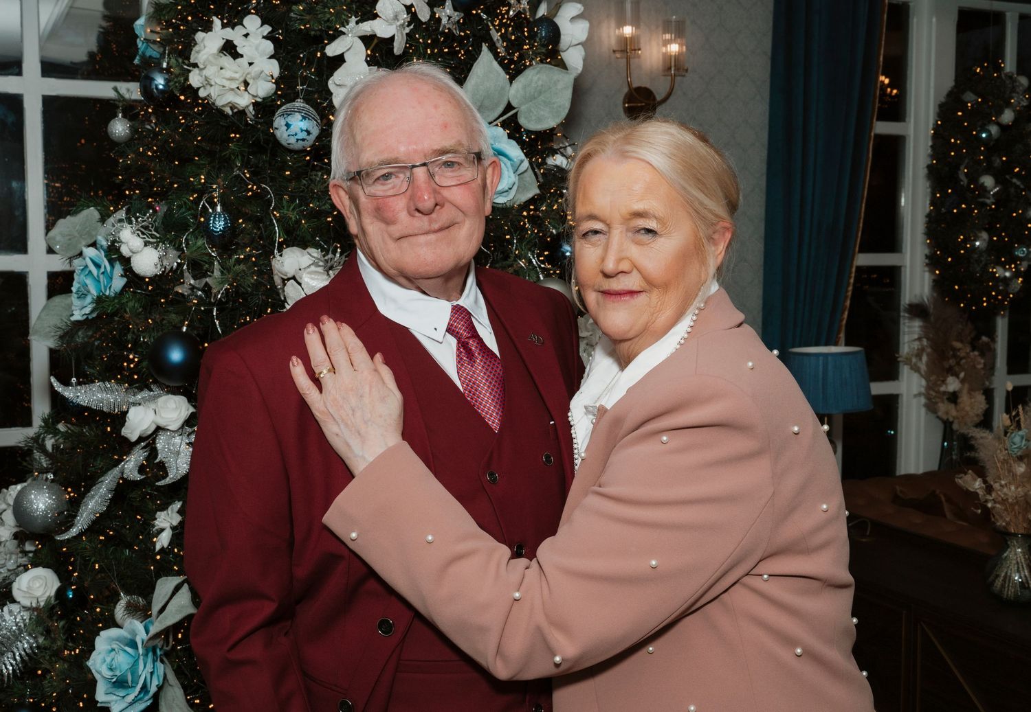 Elderly couple embracing in front of decorated Christmas tree with white and blue ornaments.
