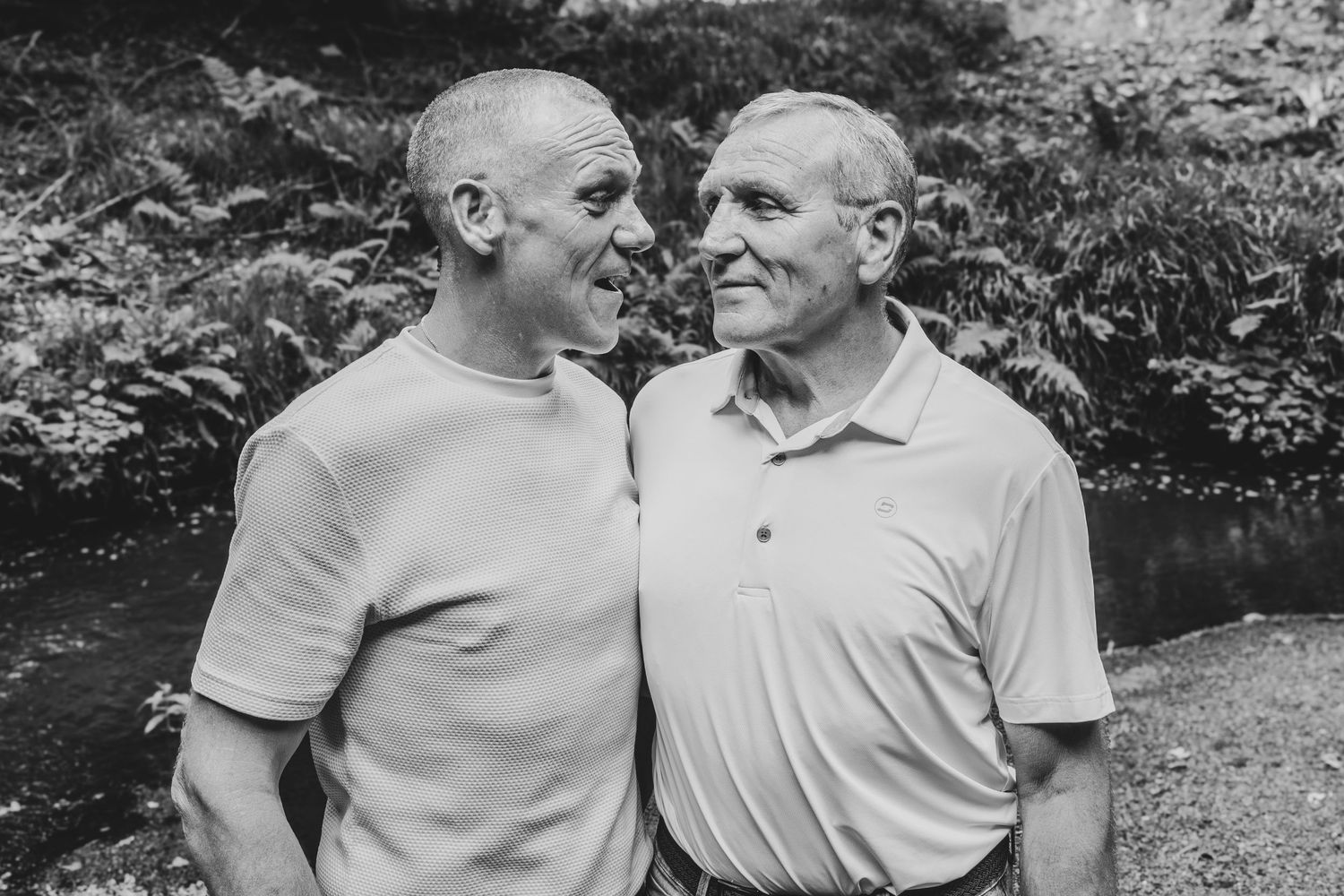 Two elderly men share a warm smile outdoors, surrounded by lush greenery, in a black and white portrait photo.
