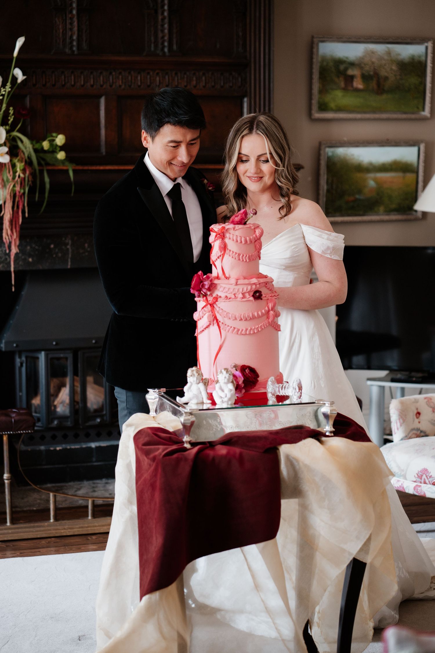 Bride and groom admire an elegant pink tiered wedding cake with floral details on a decorated table.