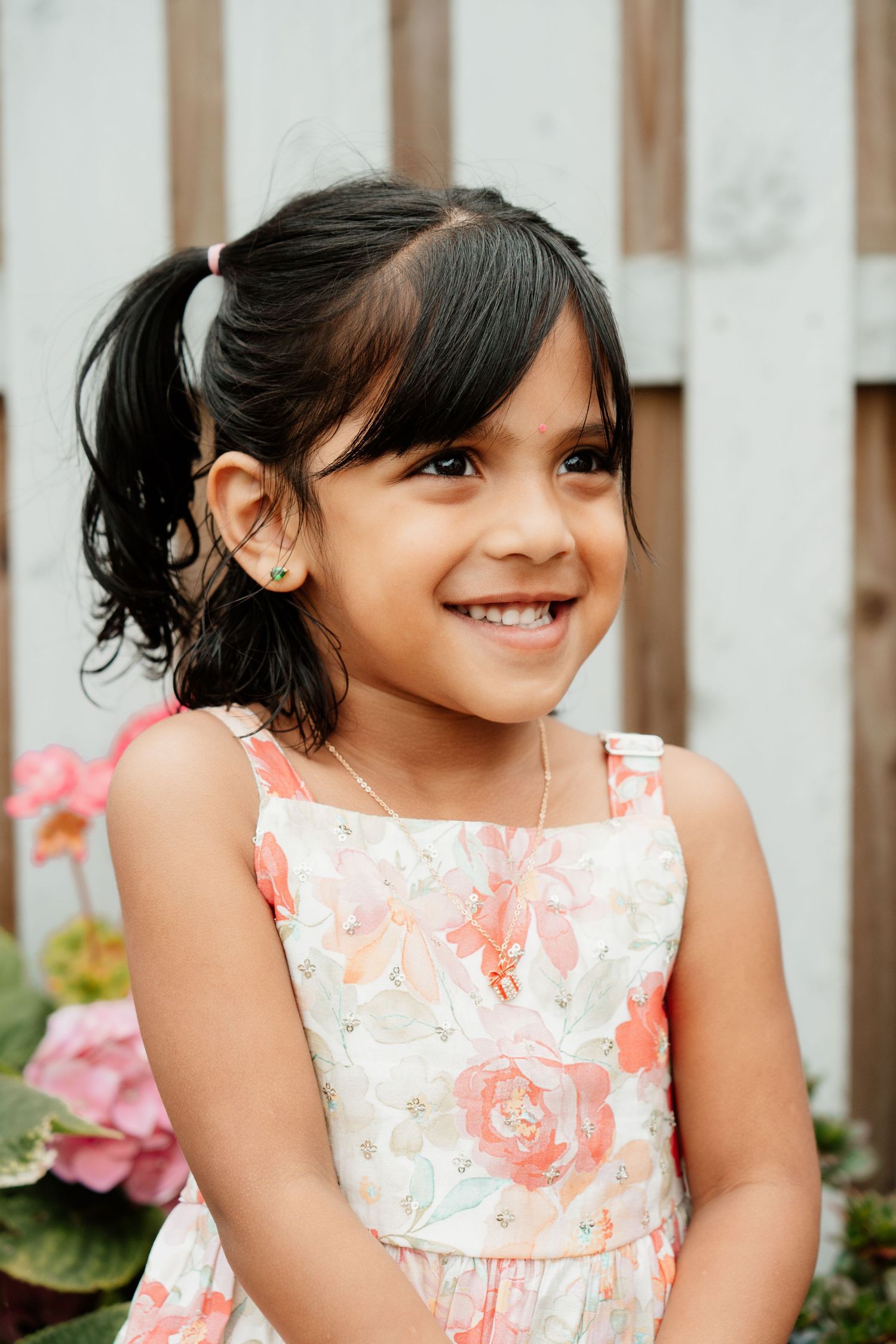 A smiling young girl with pigtails wearing a floral dress and gold earrings, posing outdoors near pink flowers.