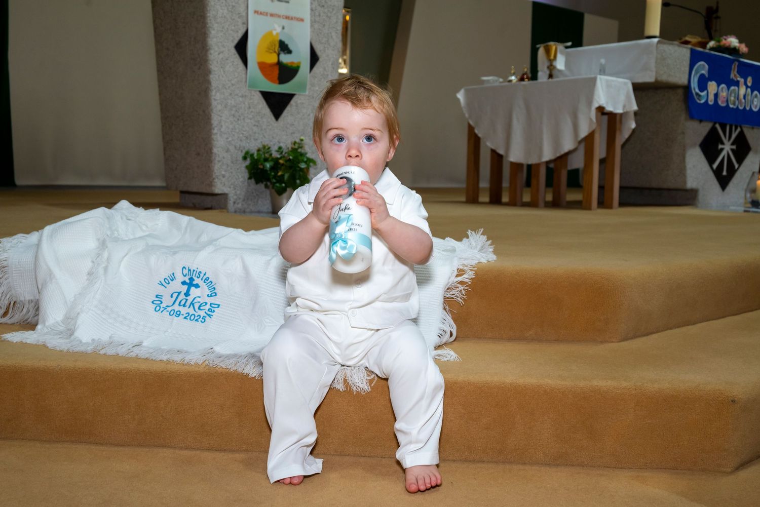 Baby in white outfit sitting on church steps, drinking from a bottle during a baptism ceremony.