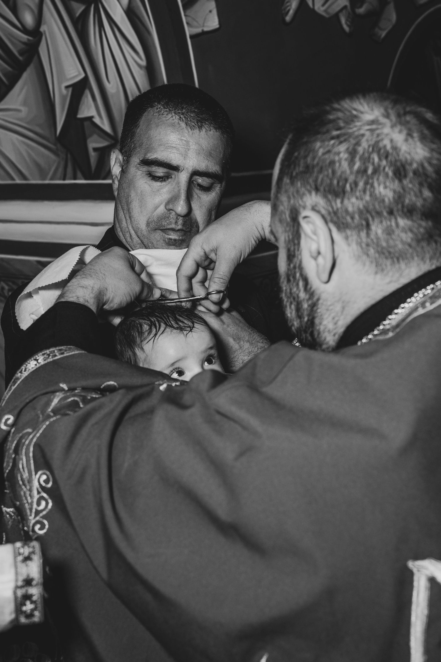 Black and white photo of a priest performing a baptism ceremony on an infant held by a godfather in a church.