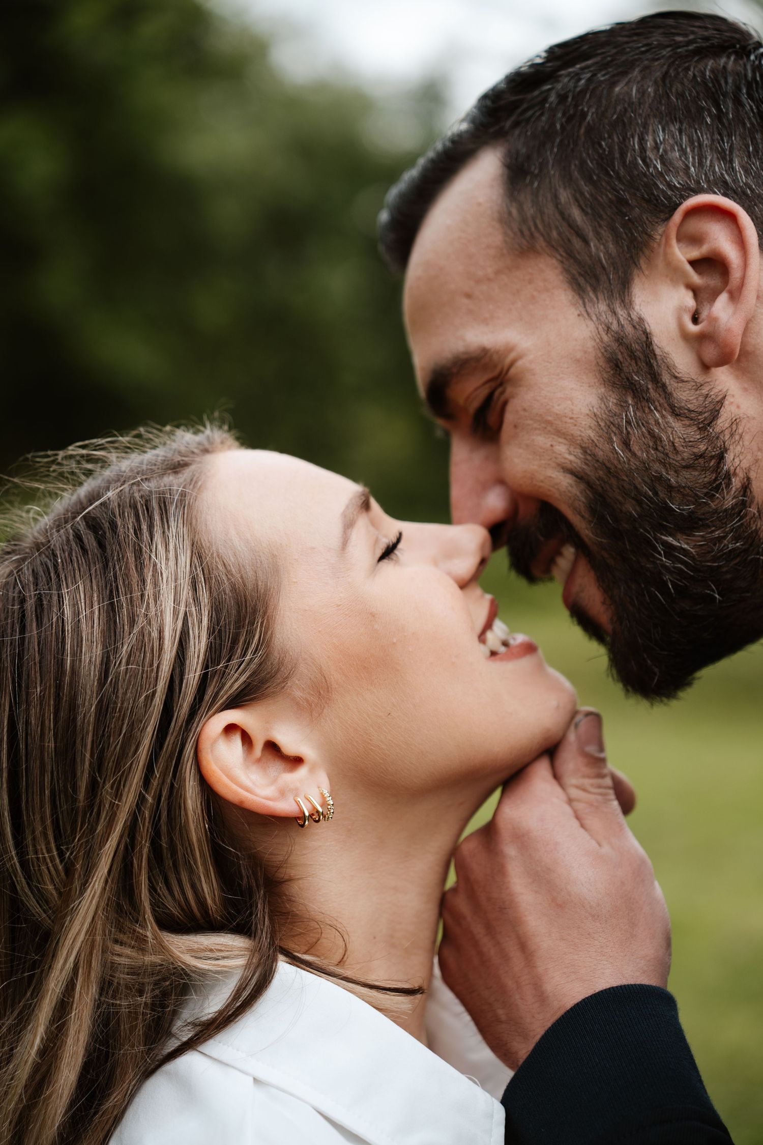 A couple shares a tender, smiling moment outdoors, the man gently holding the woman's chin as they lean in for a kiss.