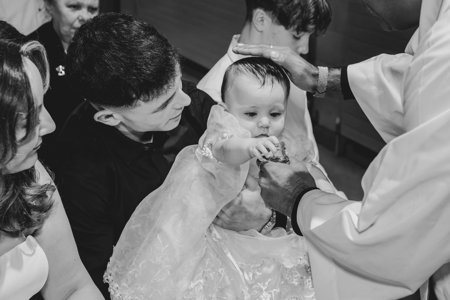 Black and white photo of a baby in white christening gown being baptized by a priest surrounded by family.