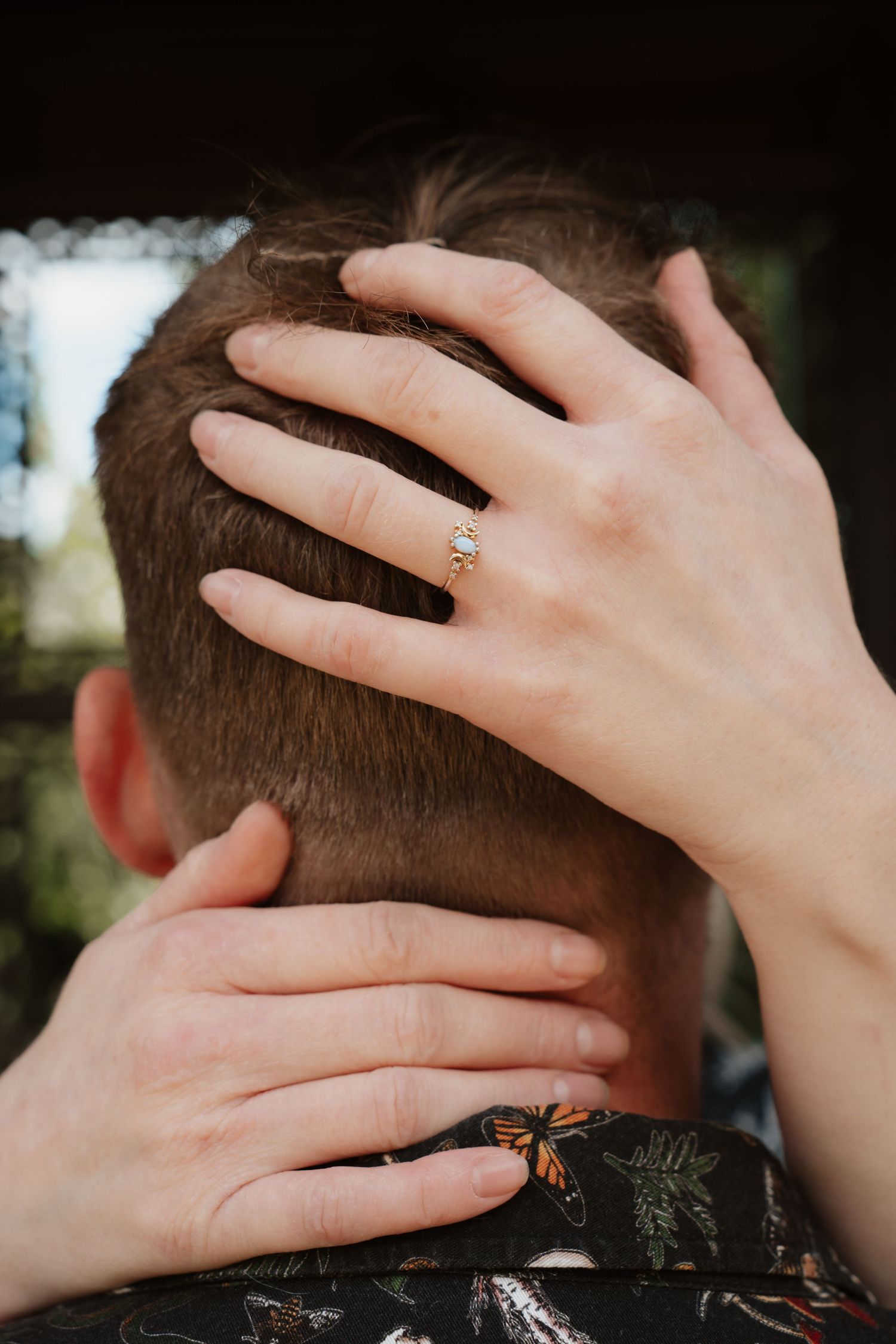 Close-up of a woman's hand with a diamond engagement ring resting on a man's head.