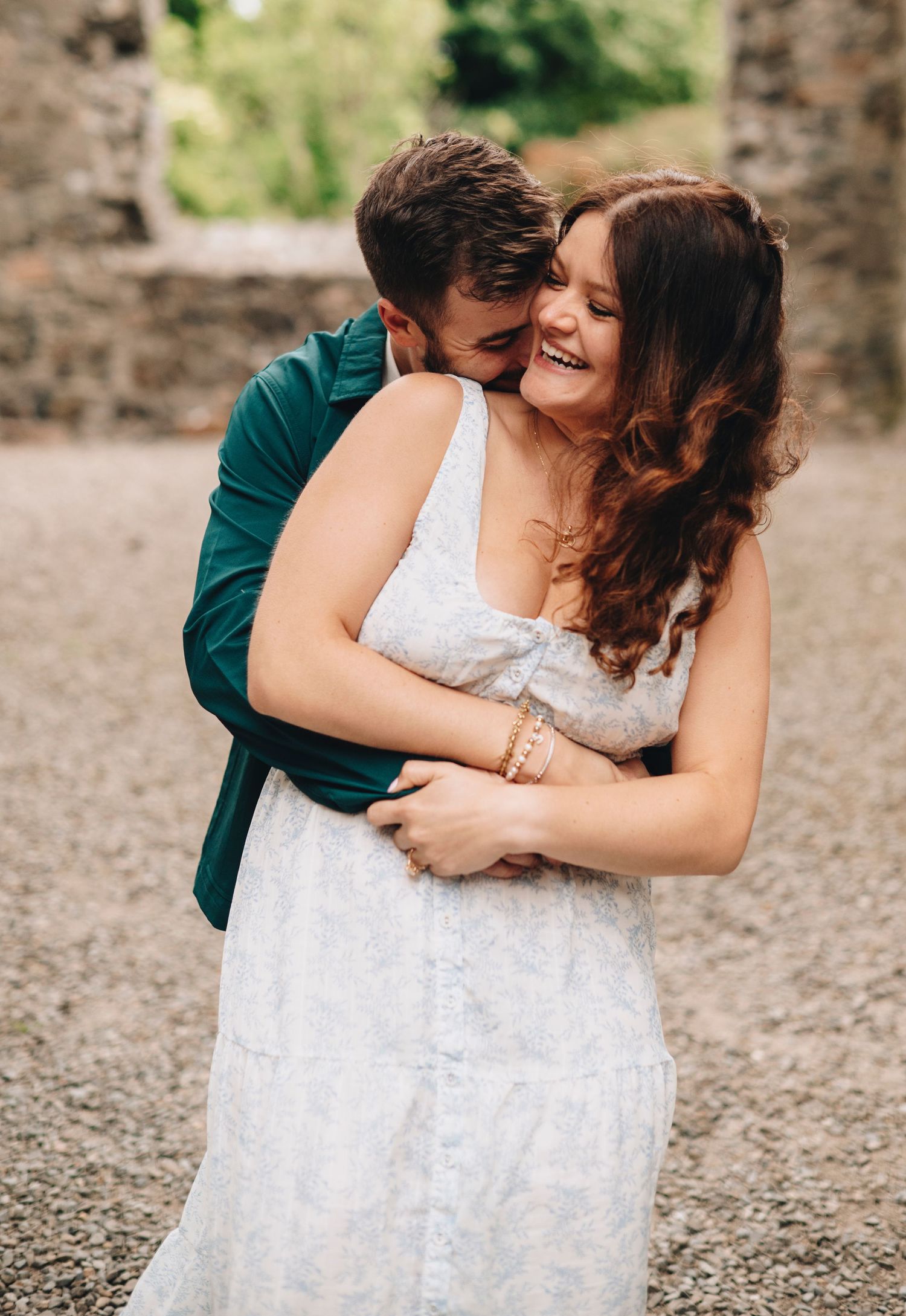 A couple embraces joyfully outdoors, woman in white dress laughing as man in green jacket hugs her from behind near stone wall.