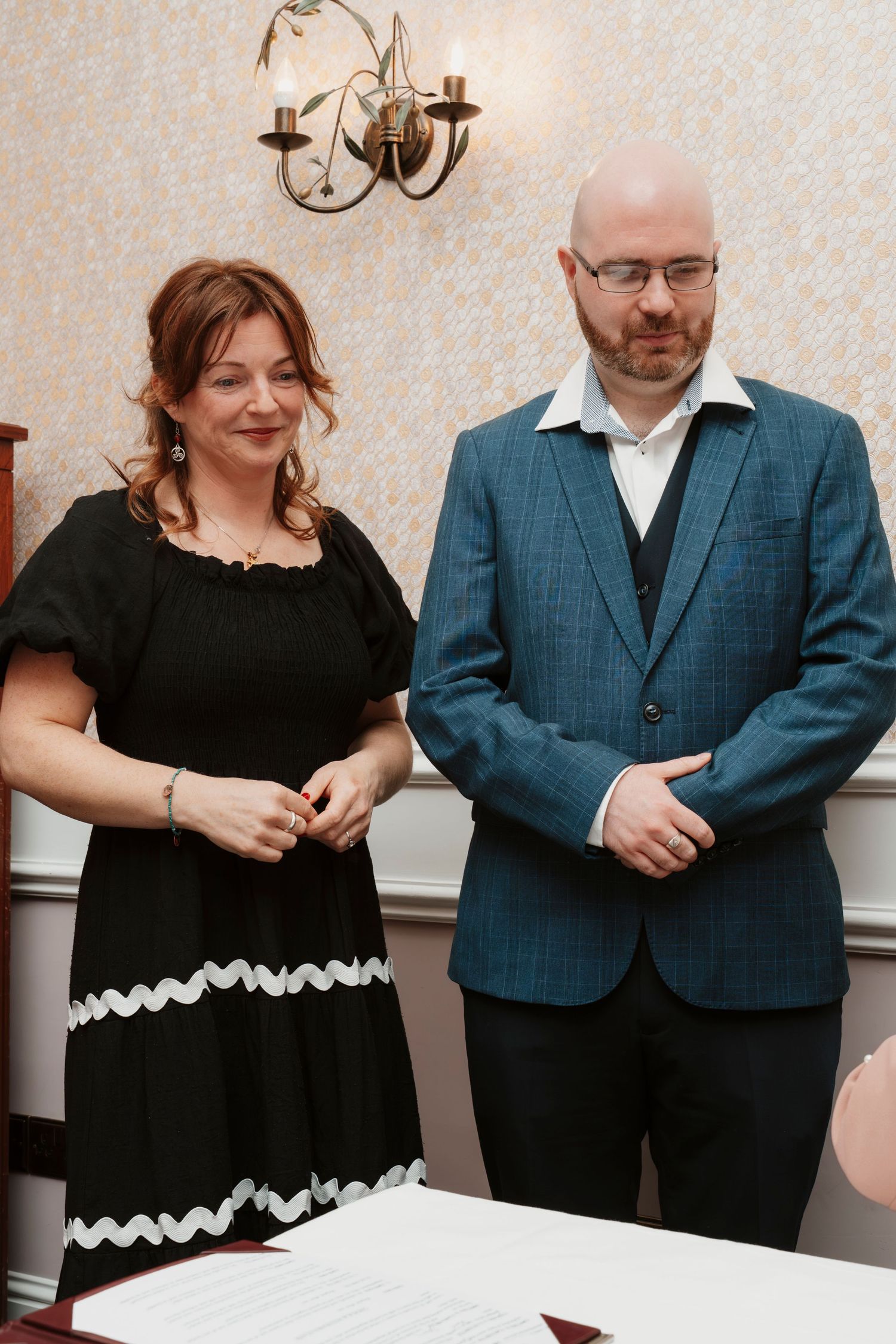 A woman in a black dress with white trim stands beside a man in a blue suit jacket in a formal interior setting.