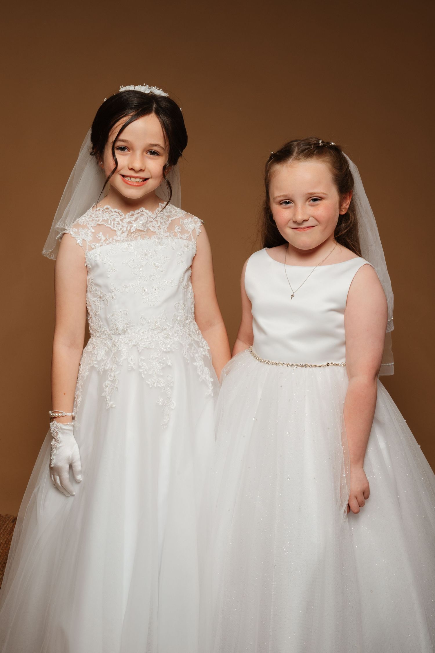 Two young girls in white First Communion dresses and veils pose together smiling for a formal portrait photo.