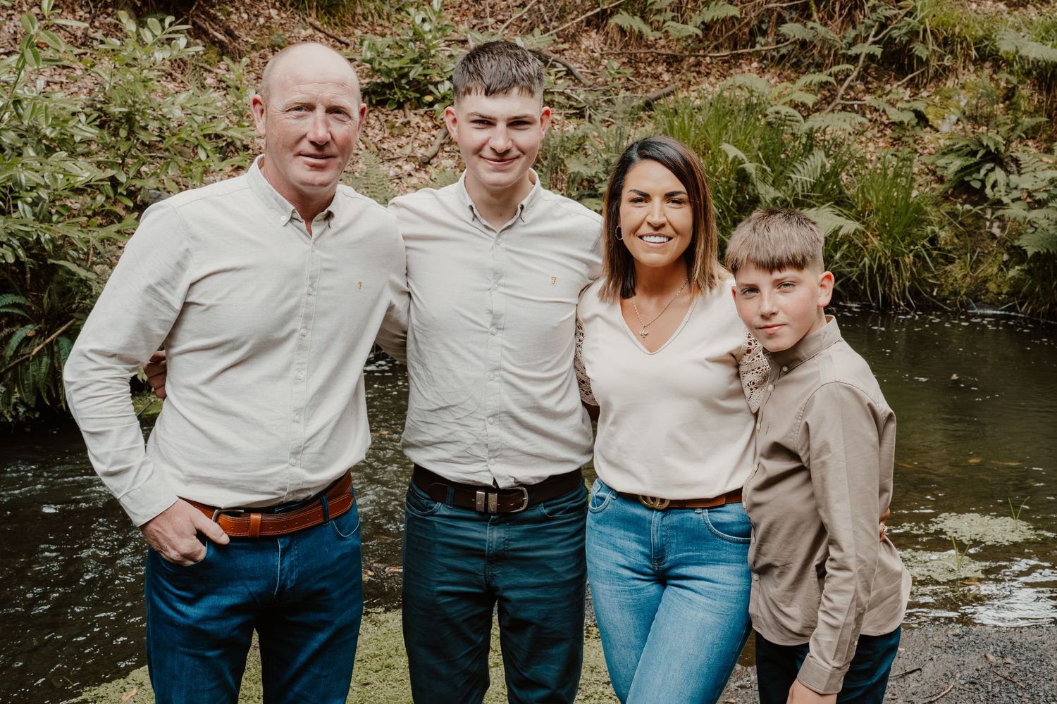 Family of four smiling outdoors near a pond surrounded by lush green ferns and trees.