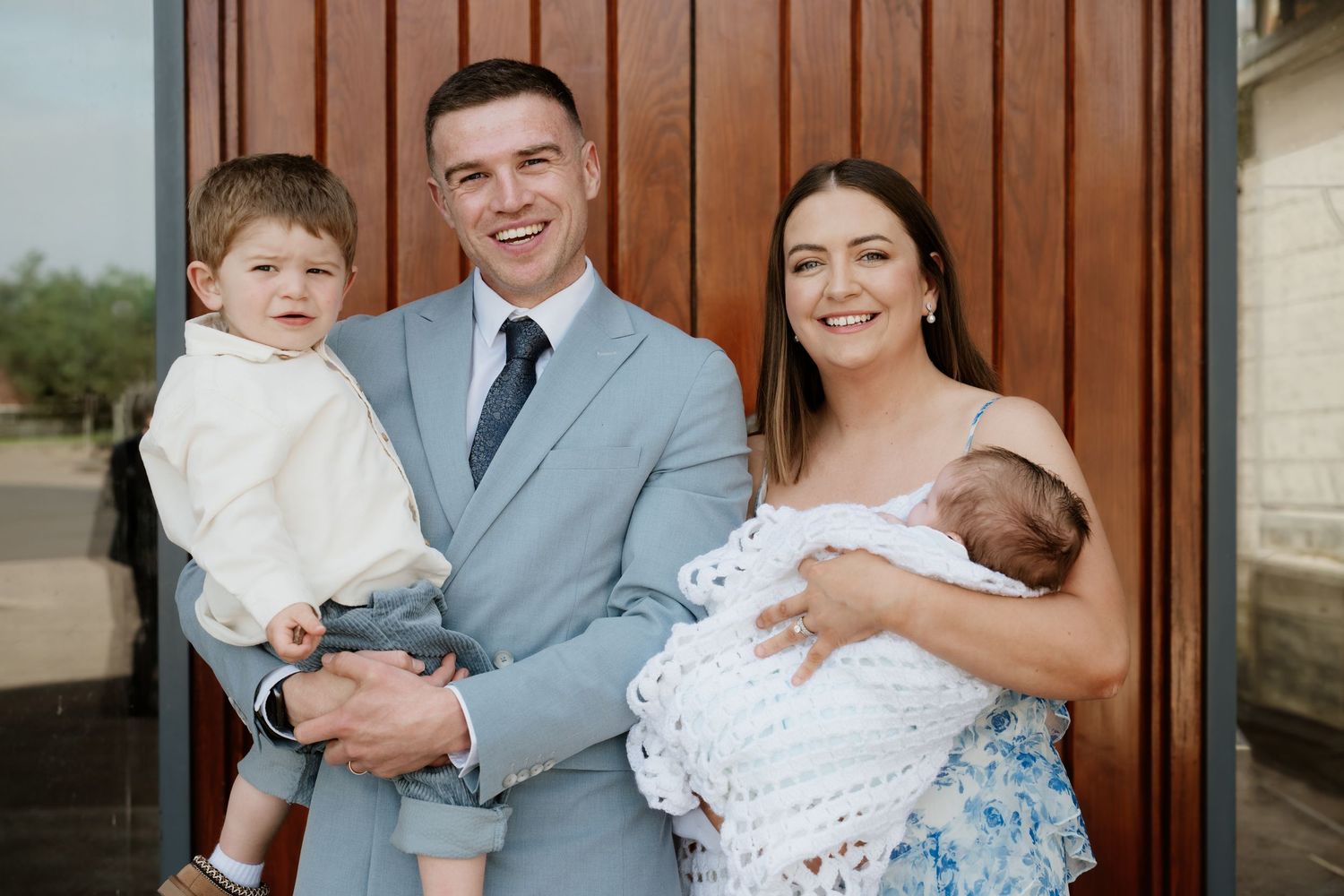 Family of four posing outdoors, parents holding two young children, dressed in formal attire for a special occasion.