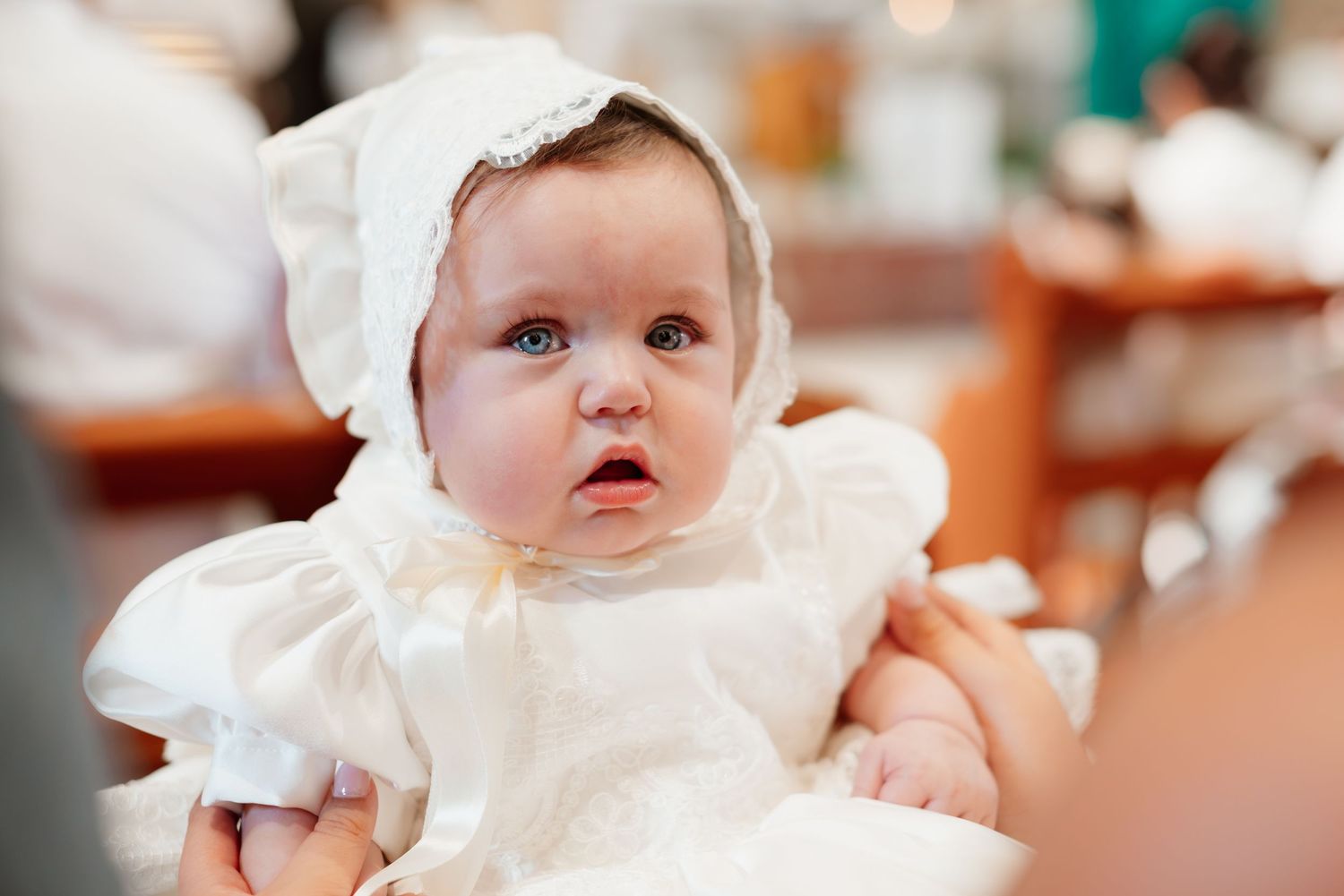 Adorable baby in white christening gown and bonnet with blue eyes at a baptism ceremony.