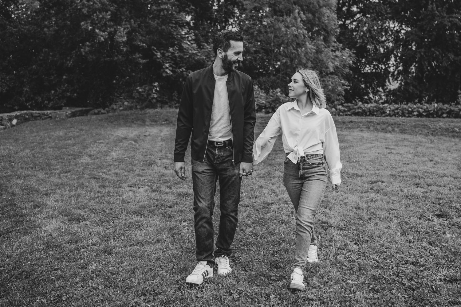 Black and white photo of a couple holding hands and walking together on a grassy lawn surrounded by trees.