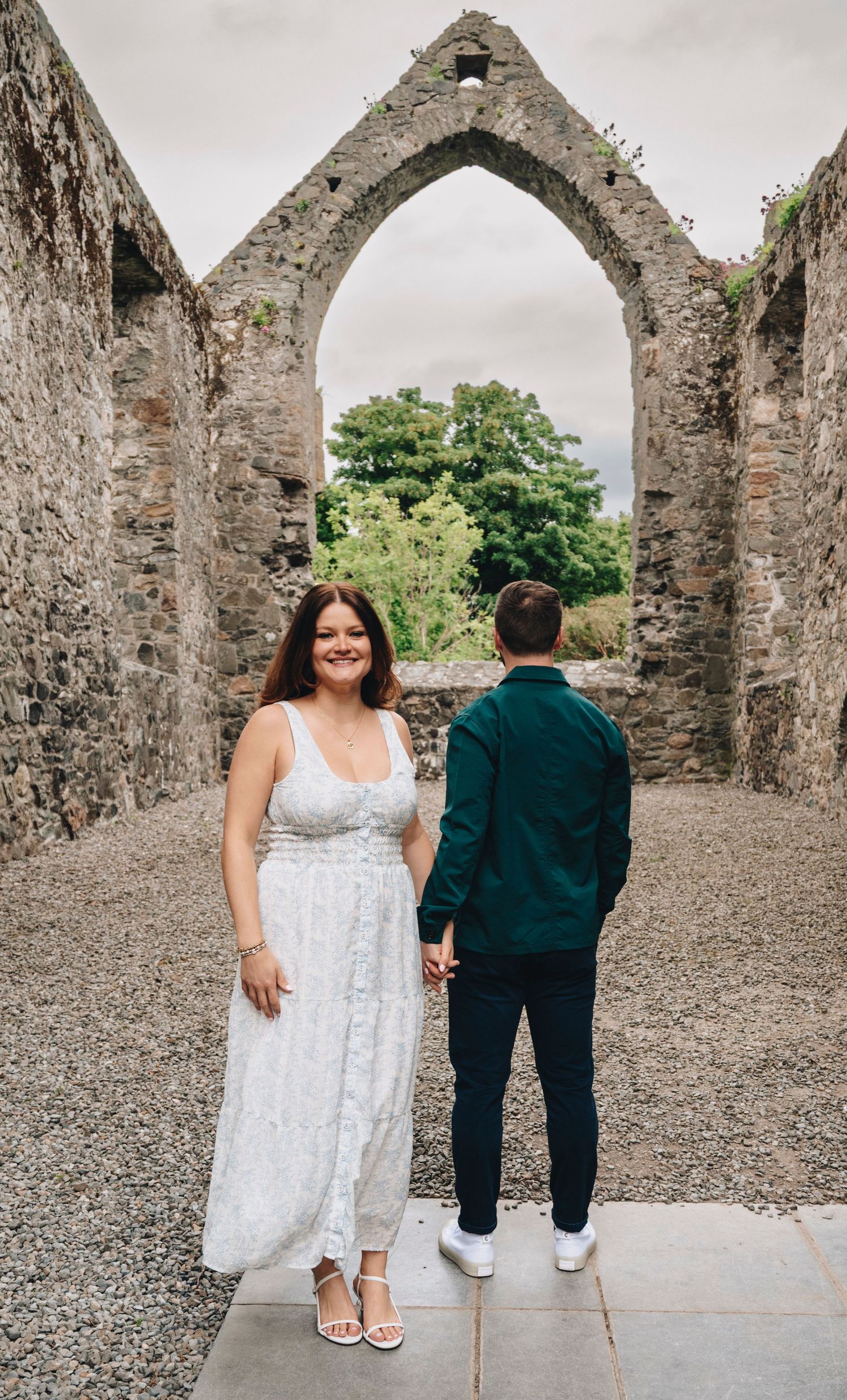 Couple holding hands at ancient stone ruins, woman in white dress smiling at camera, man in teal jacket facing away.