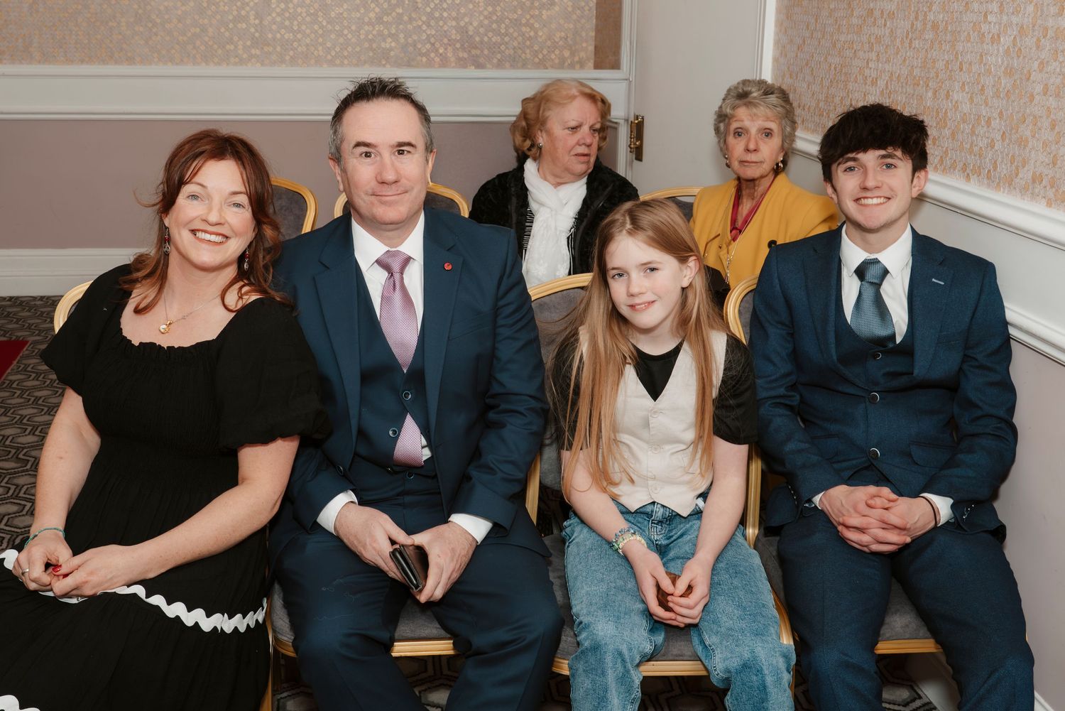 Family portrait of six people in formal attire seated together on a bench in an elegant indoor setting.