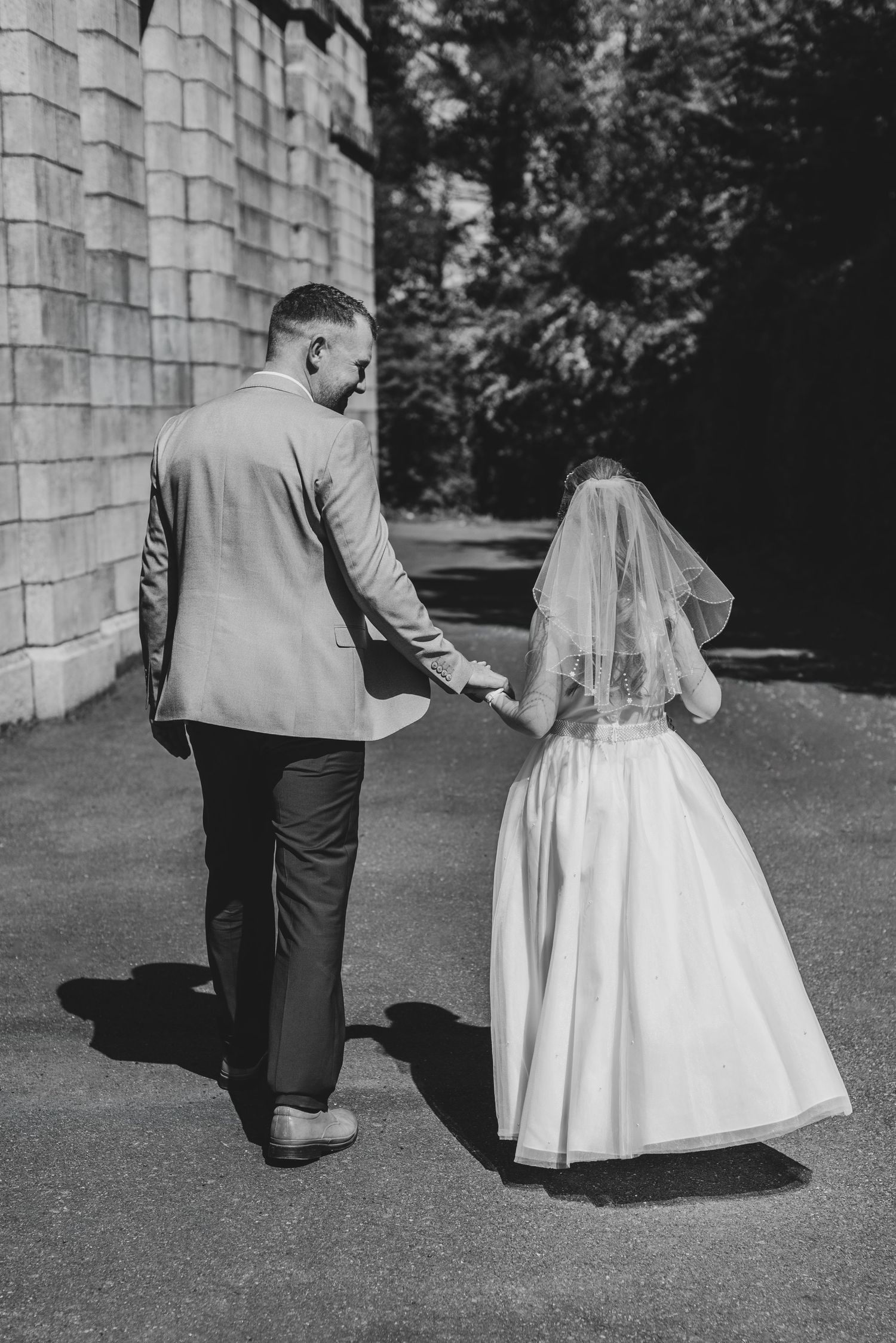 Black and white photo of a bride and groom walking hand in hand, viewed from behind, near a stone building.
