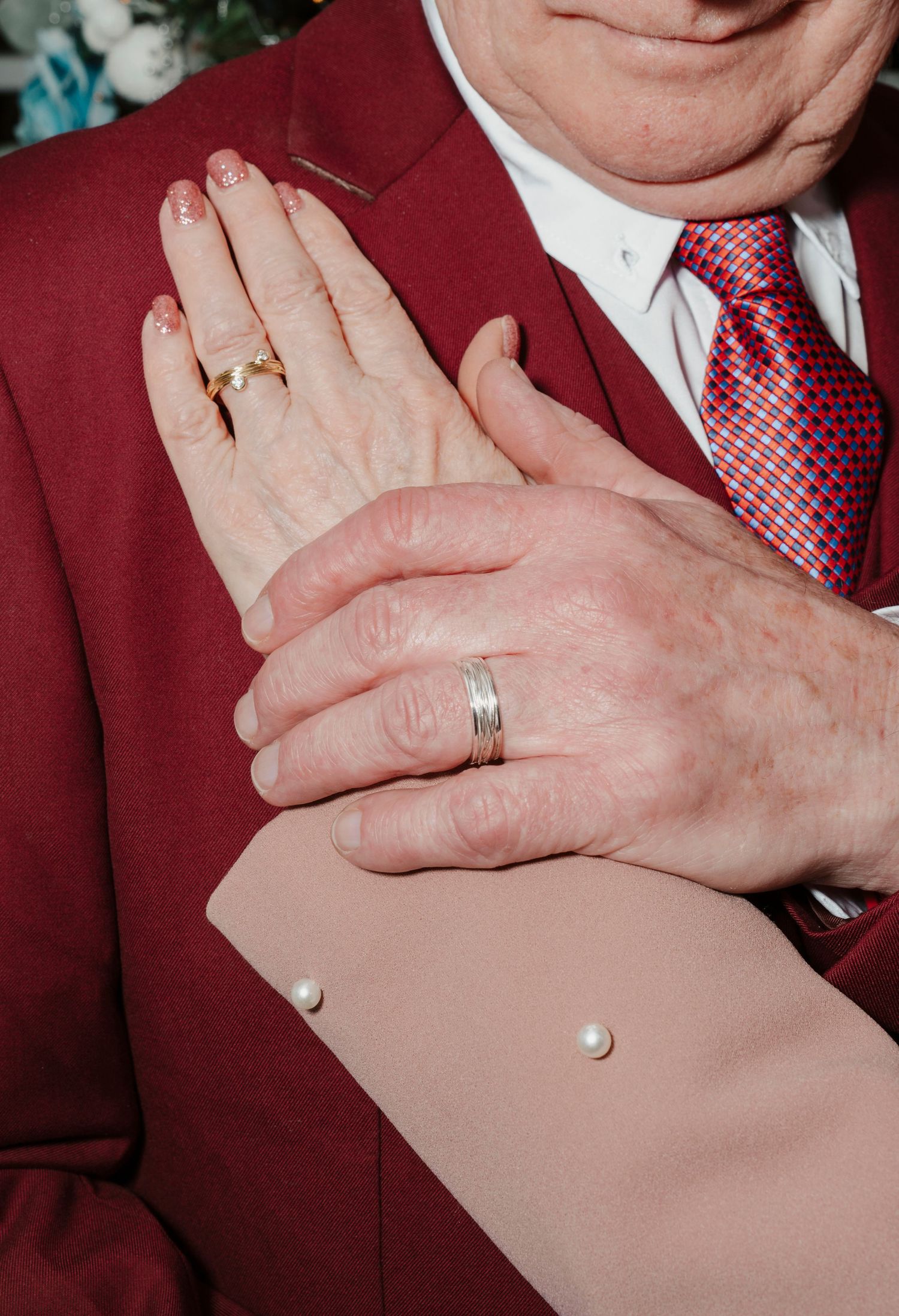 Elderly couple's hands clasped together, wearing wedding rings, man in burgundy suit with red tie, woman in pearl-buttoned outfit.