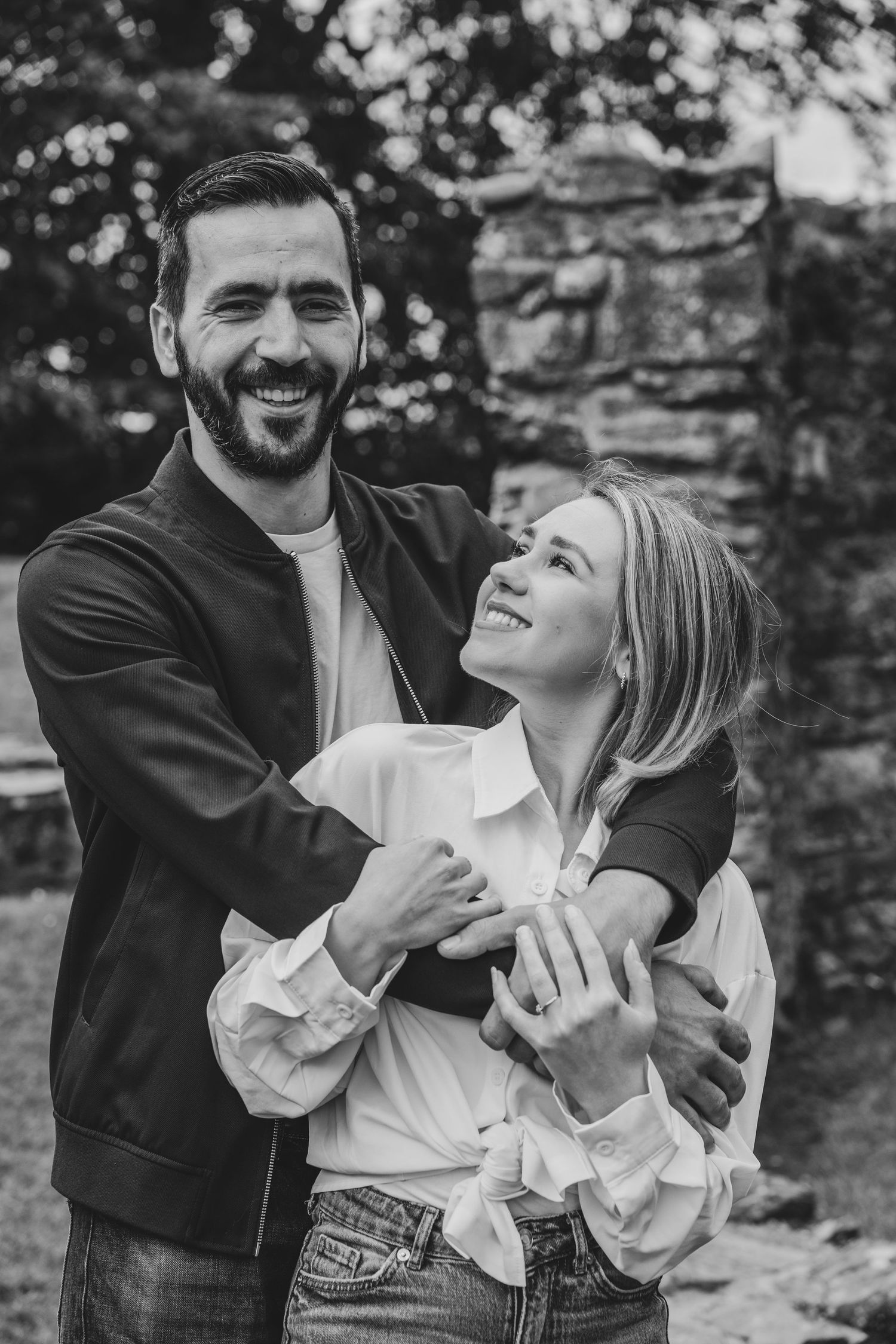 Black and white photo of a smiling couple embracing outdoors, man in jacket hugging woman in white blouse.
