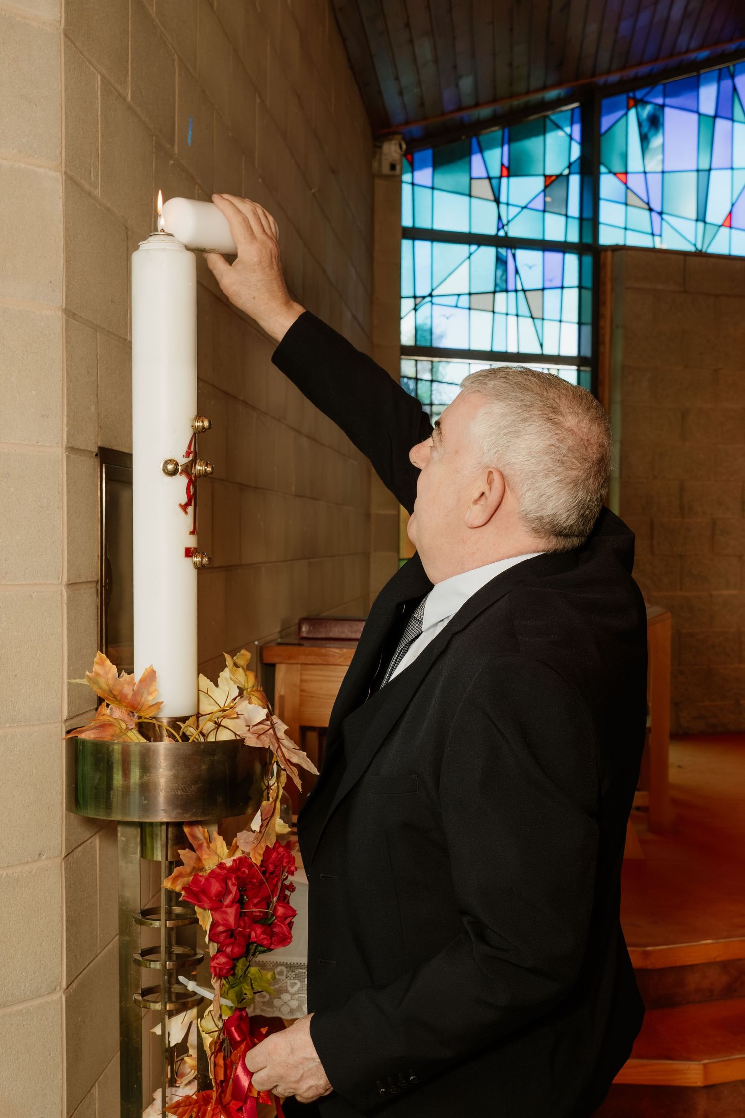 Man in black suit lighting a large white candle in a church with stained glass windows and red floral decorations.