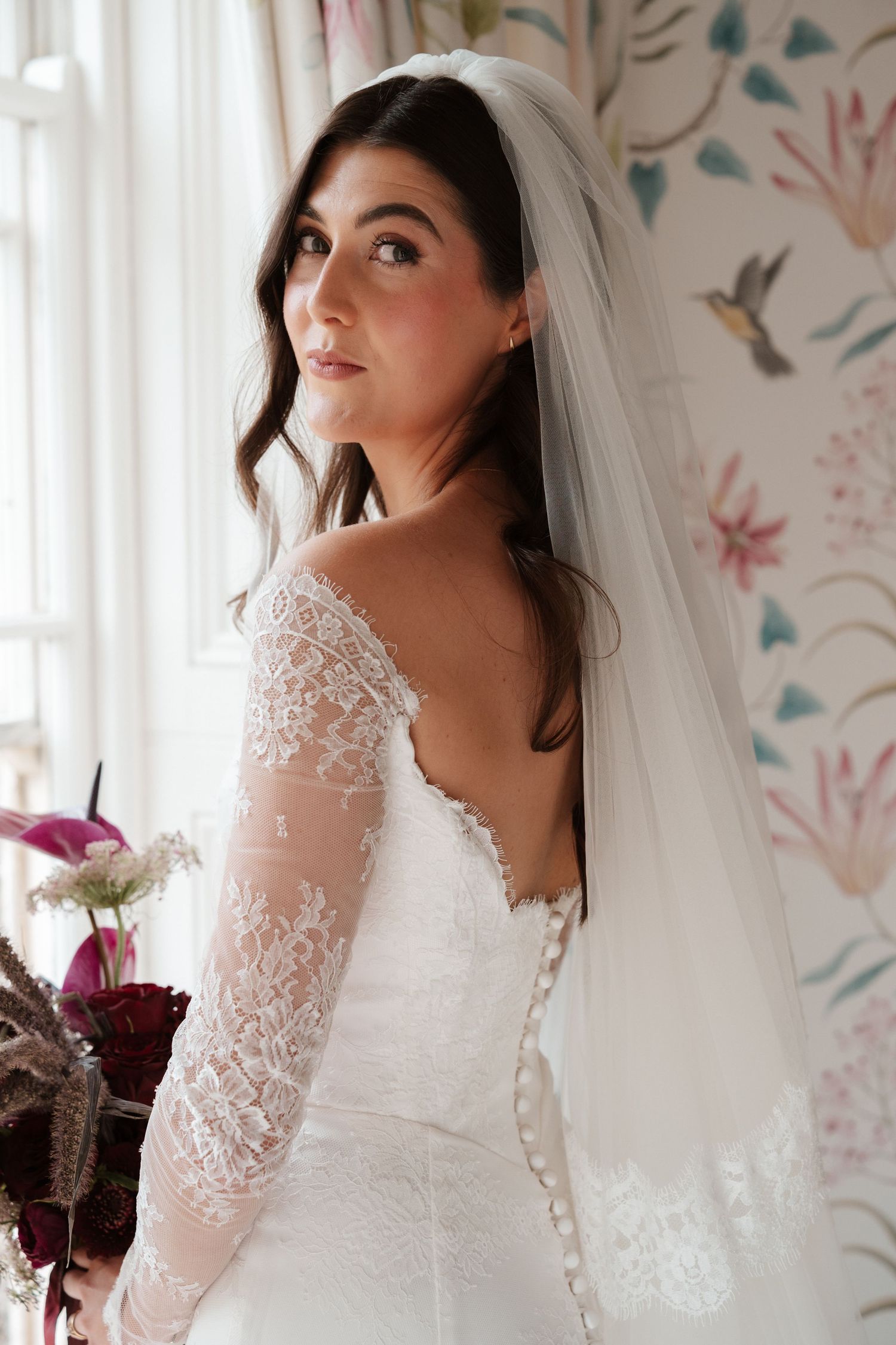 Bride in lace long-sleeve wedding gown and veil gazes over shoulder near floral wallpaper window.