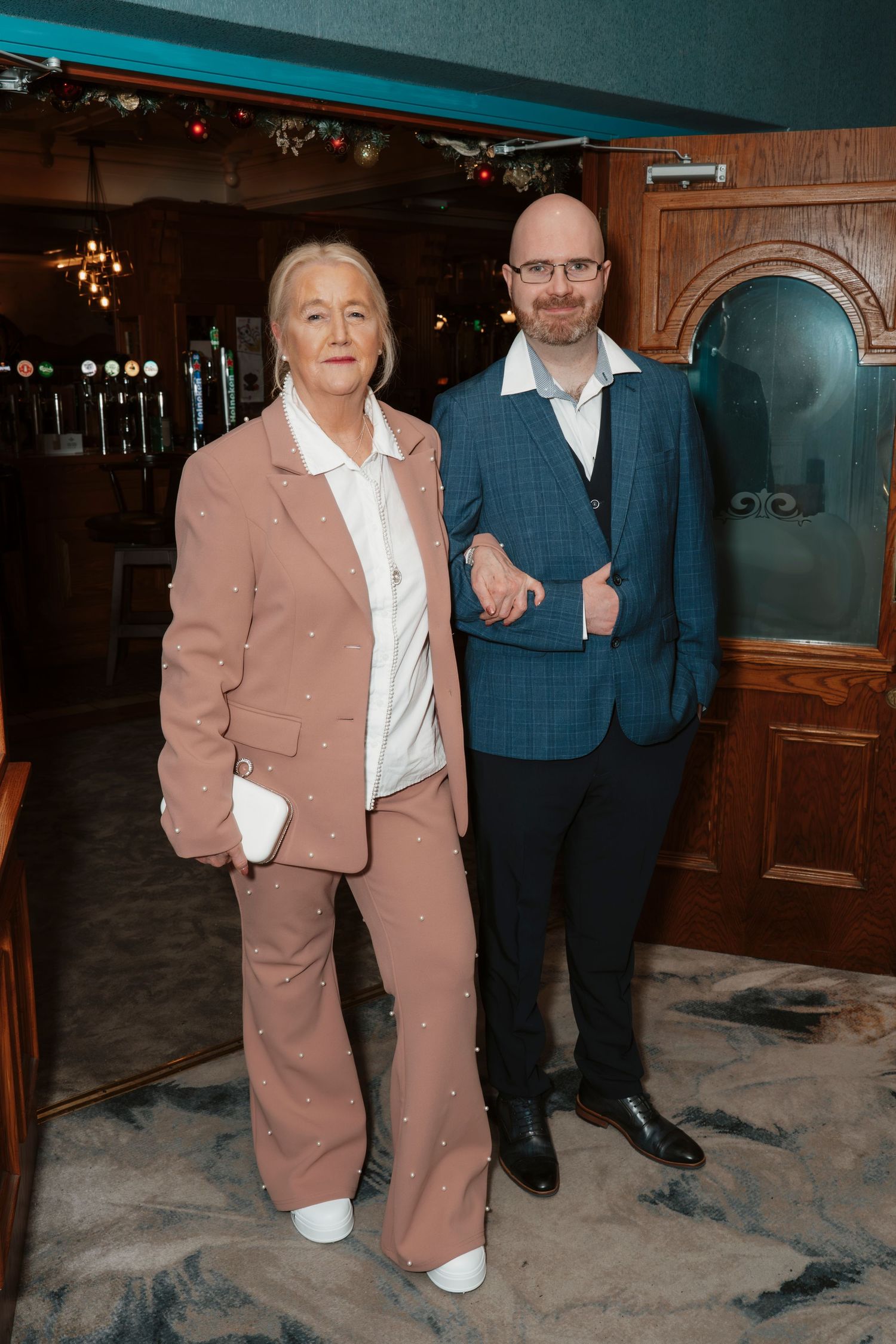 Two people in formal attire pose together in an elegantly decorated venue with vintage decor and warm lighting.