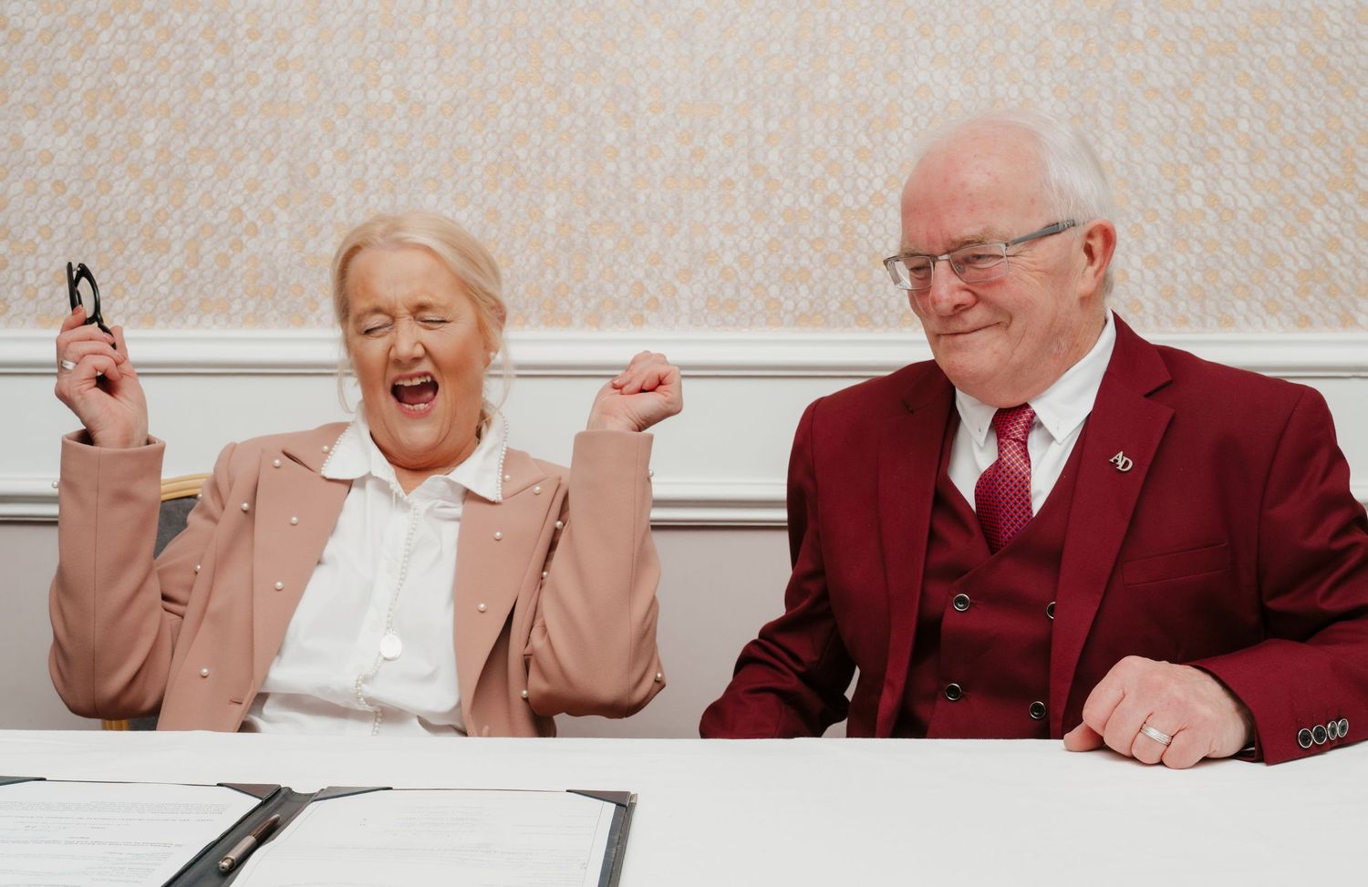 Elderly couple laughing joyfully at a table, woman raising fists in excitement, man in burgundy suit smiling.