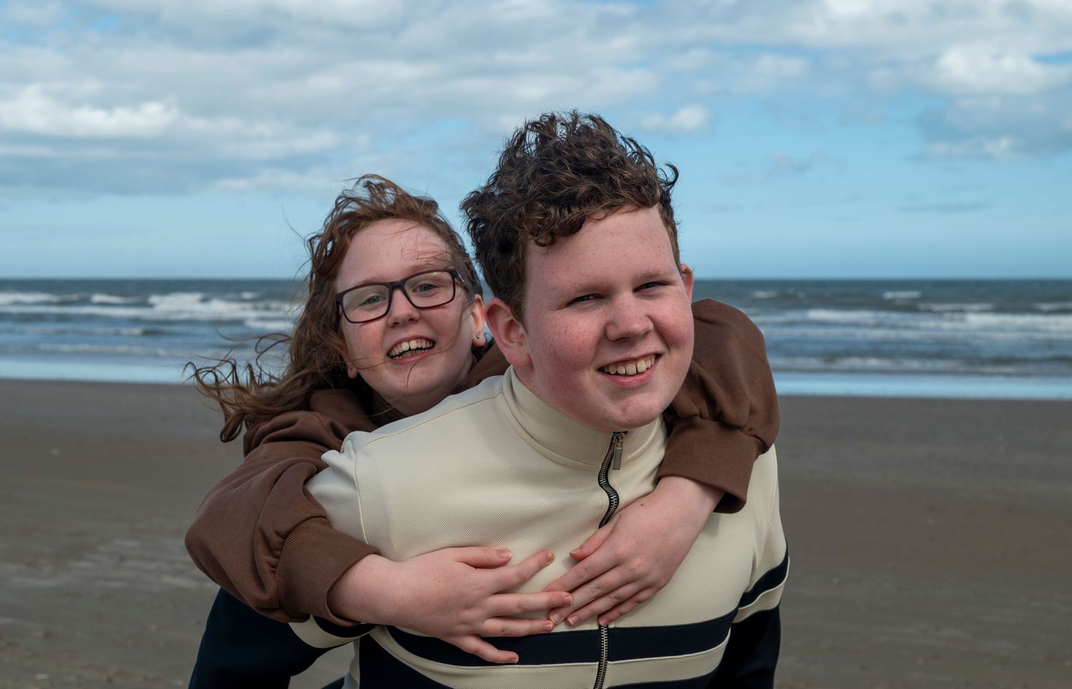 Two smiling children hugging on a sandy beach with ocean waves and cloudy sky in the background.