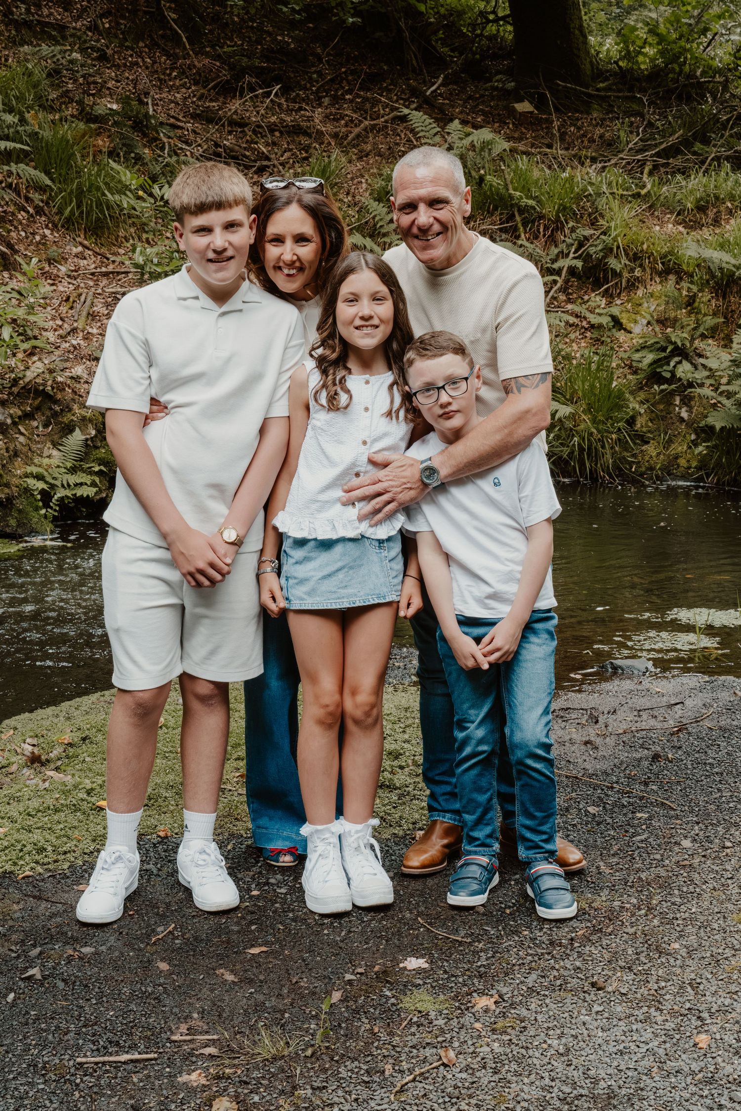 Happy family of five posing outdoors near a pond, surrounded by lush greenery, wearing casual neutral outfits.