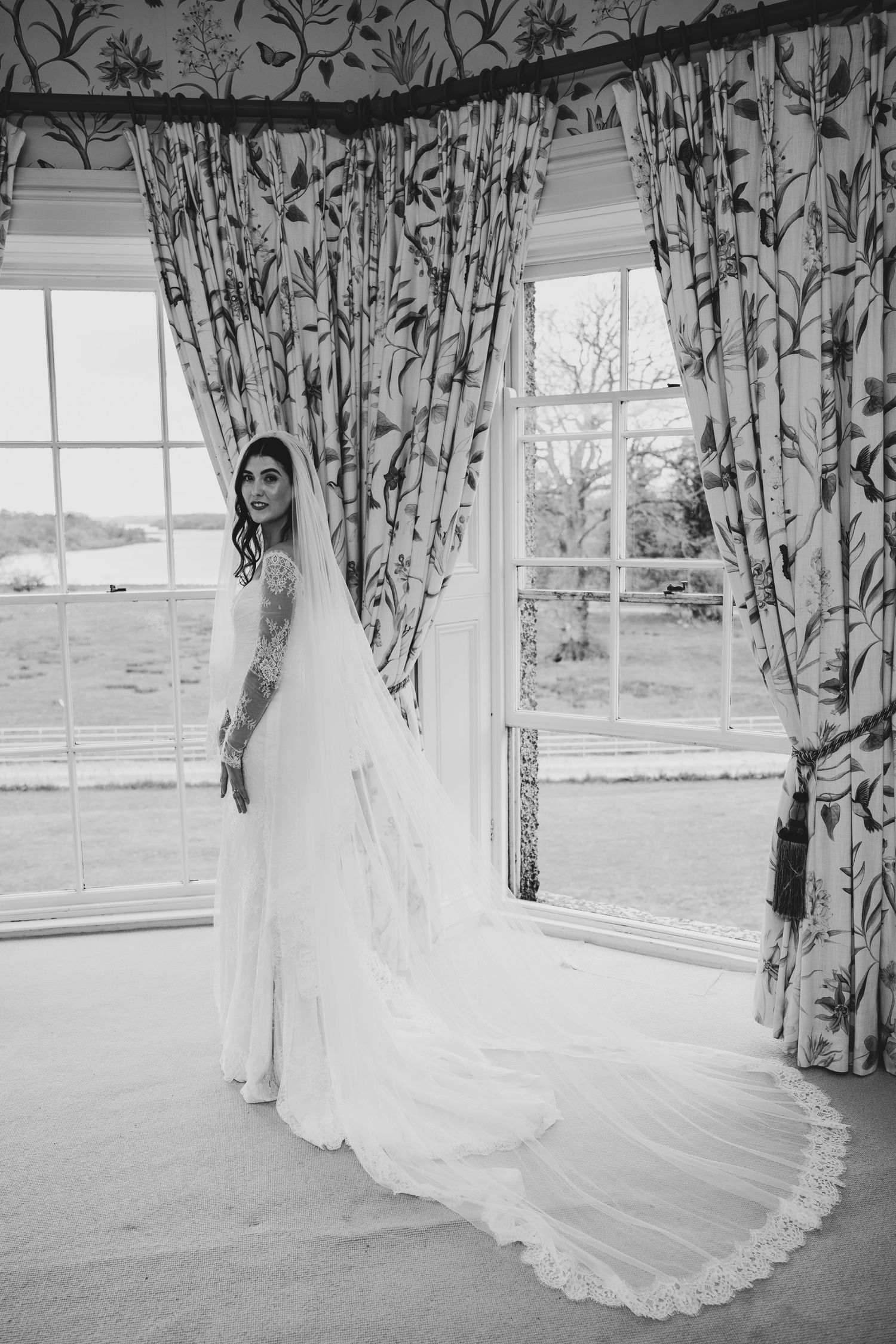 Black and white photo of a bride in a lace wedding gown and long veil standing by large windows with floral curtains.