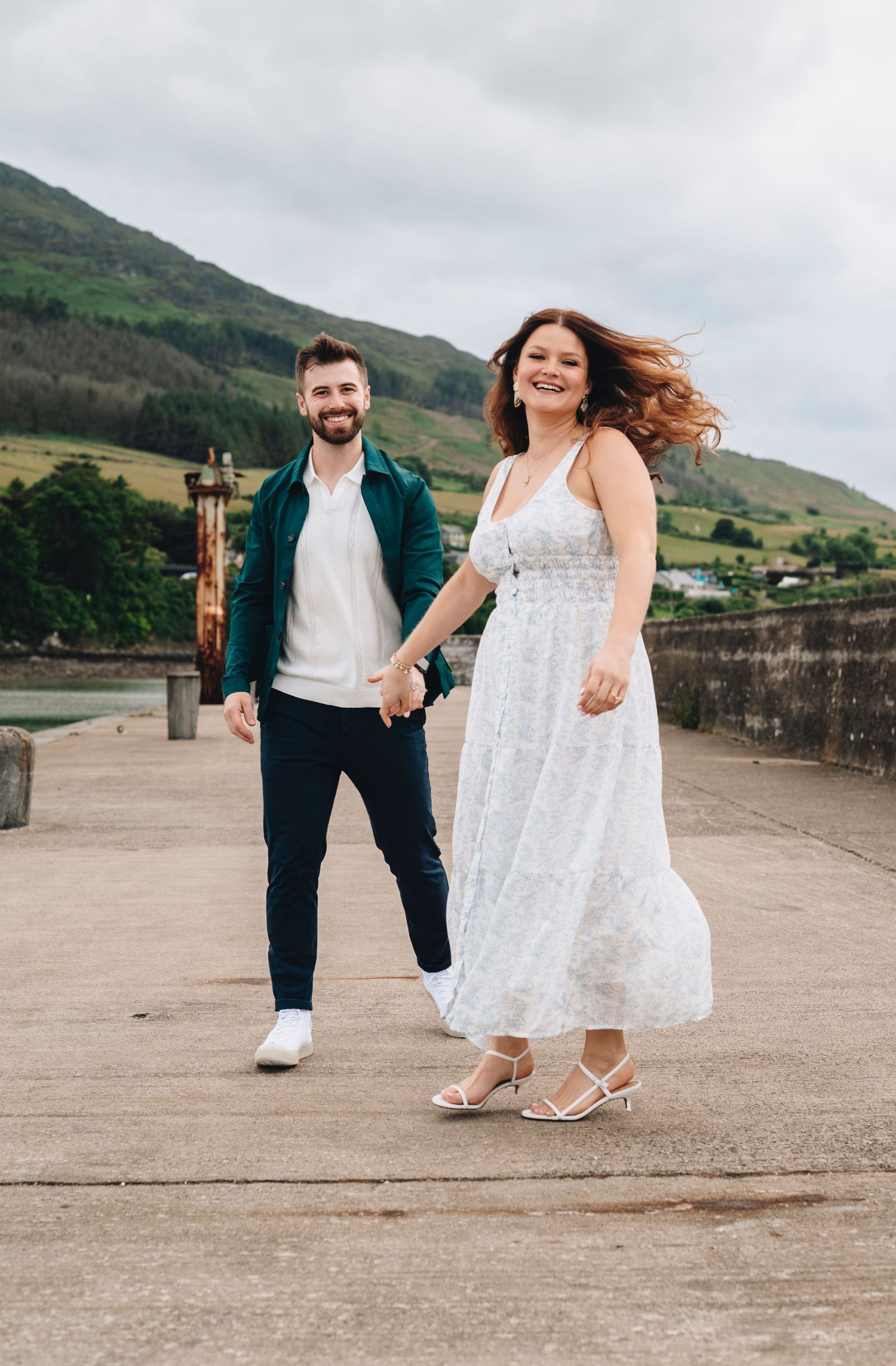 Happy couple holding hands on a seaside pier with green hills in the background.