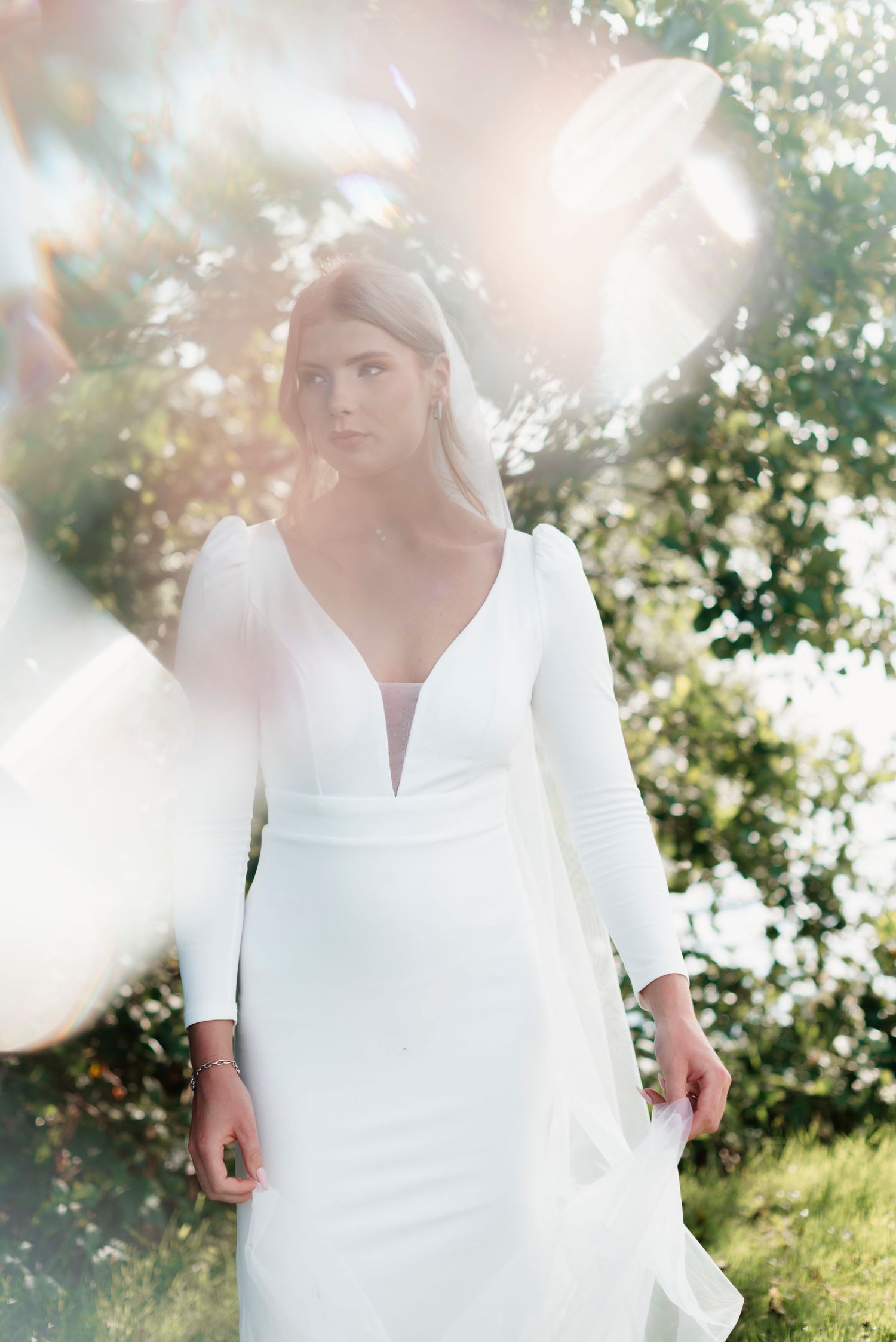 Bride in long-sleeve deep V-neck white gown stands in sunlit garden with lens flare.