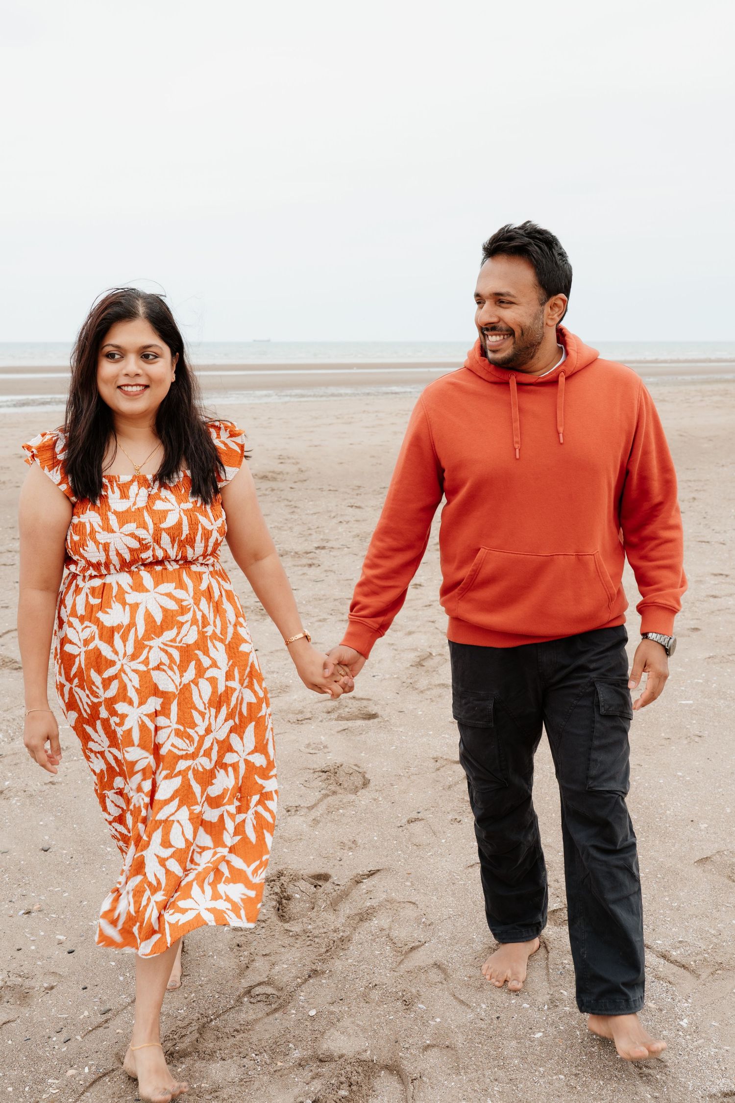 A couple walks hand-in-hand on a sandy beach, both wearing matching orange outfits, smiling joyfully.