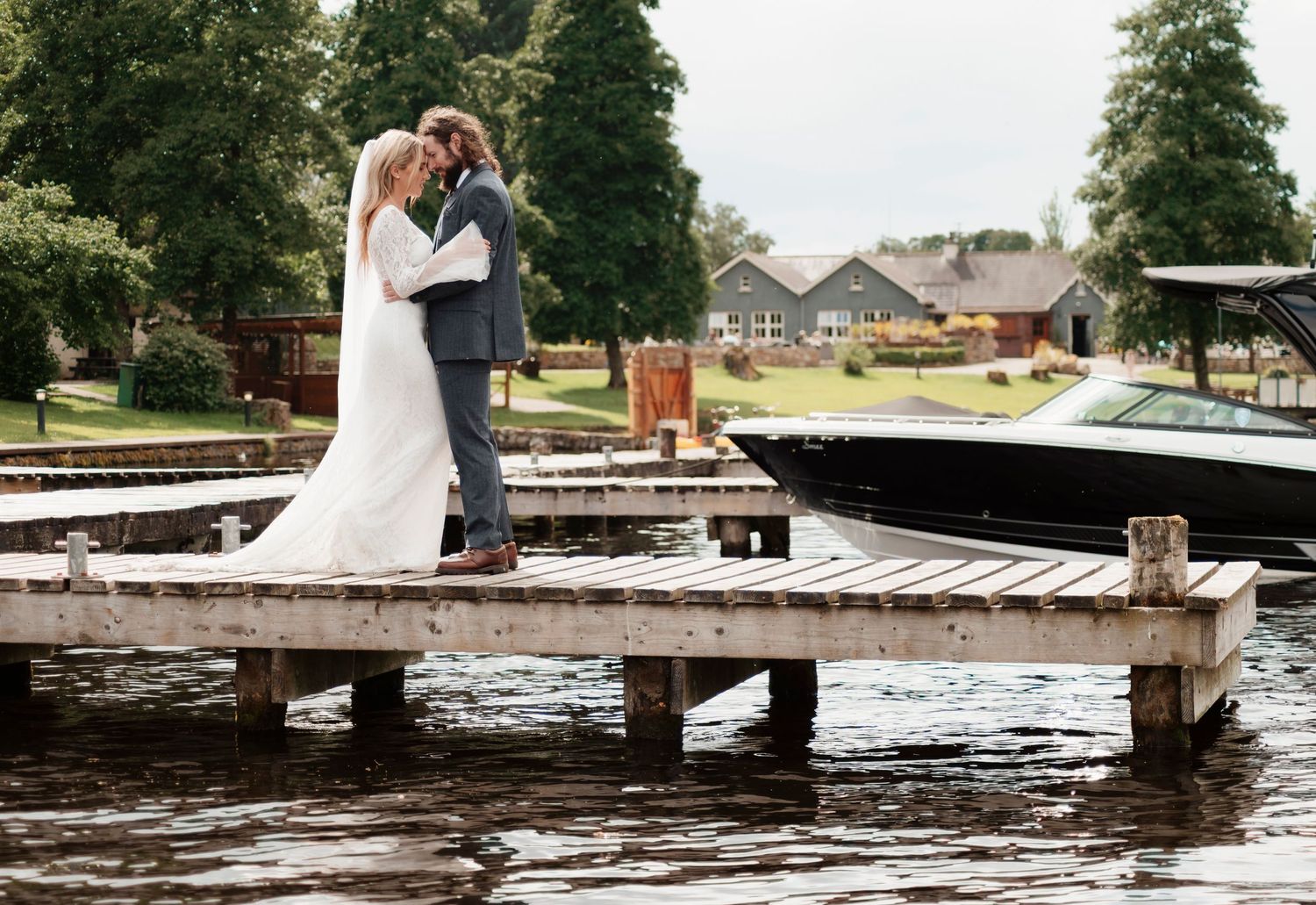 Romantic wedding couple sharing a kiss on a lakeside dock with a boat and greenery behind them.