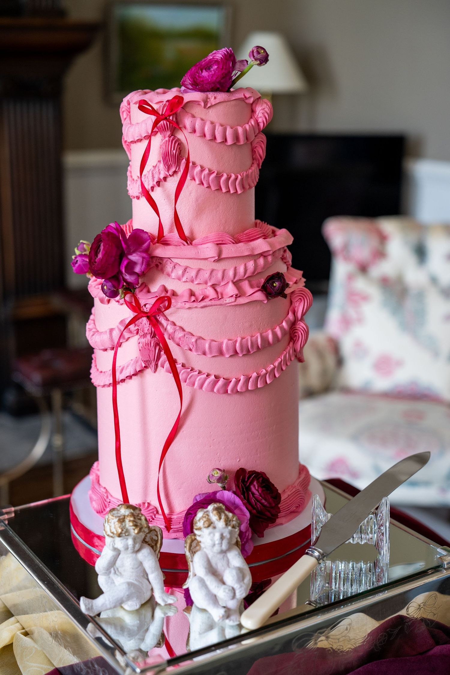 Three-tier pink fondant wedding cake decorated with ruffles, flowers, and cherub figurines on mirrored tray.