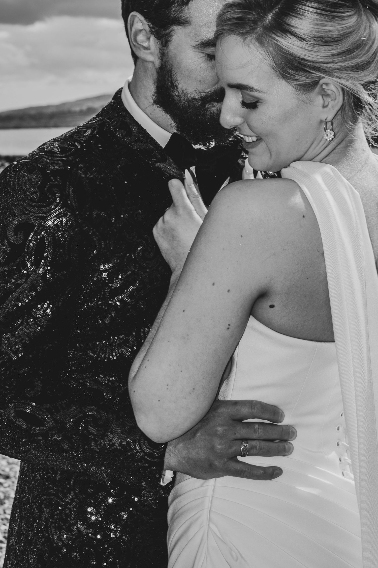 Black and white photo of a bride and groom embracing outdoors, she wears a white halter gown, he wears a sequined jacket.