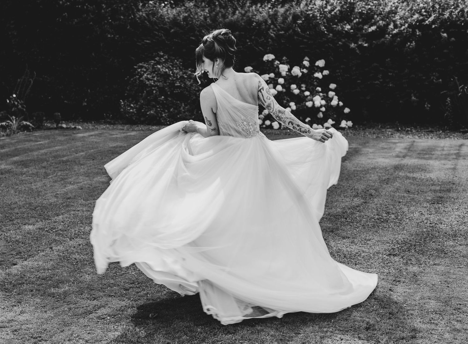 Bride twirling in elegant white wedding gown on garden lawn in black and white photography.
