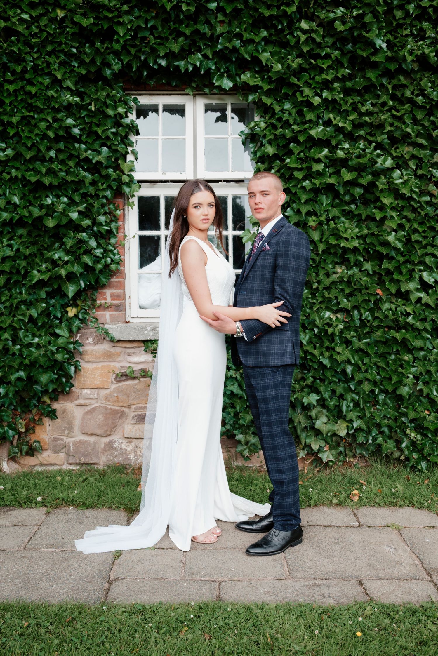 Bride in sleek white gown with veil poses with groom in plaid suit before ivy-covered stone wall.