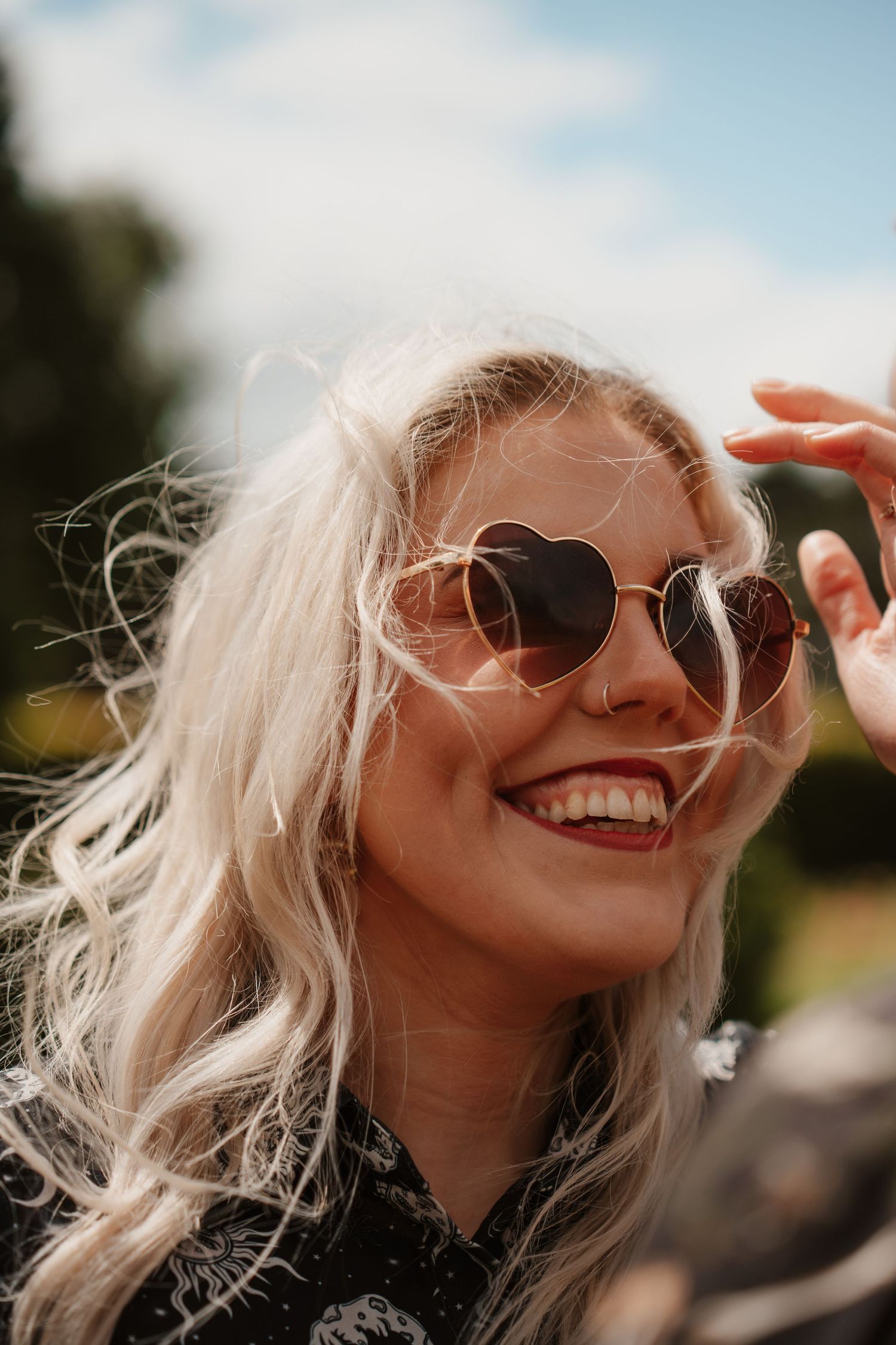 Smiling blonde woman wearing heart-shaped sunglasses outdoors on a sunny day.