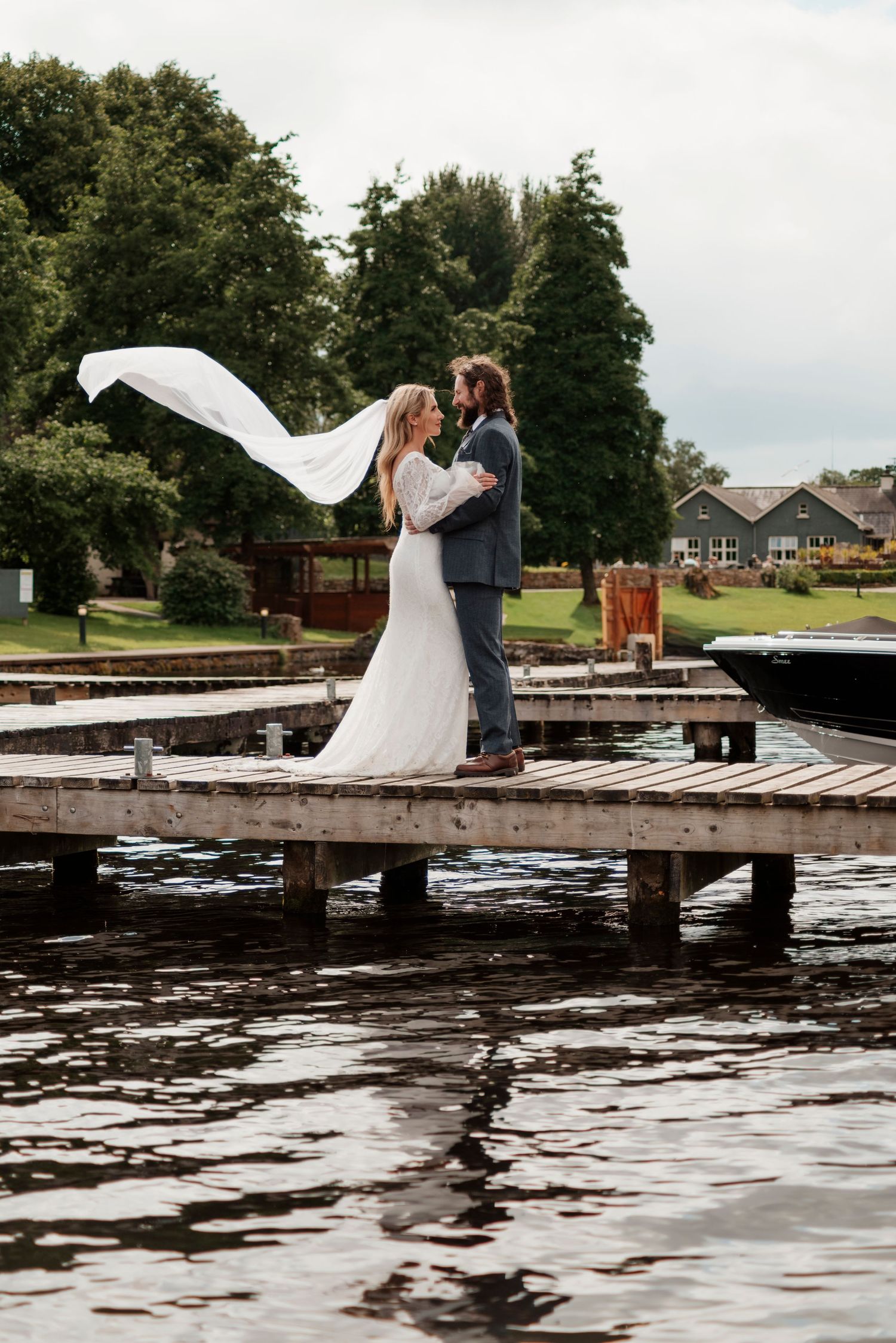 Bride and groom embrace on wooden dock by lake, veil flowing in wind.
