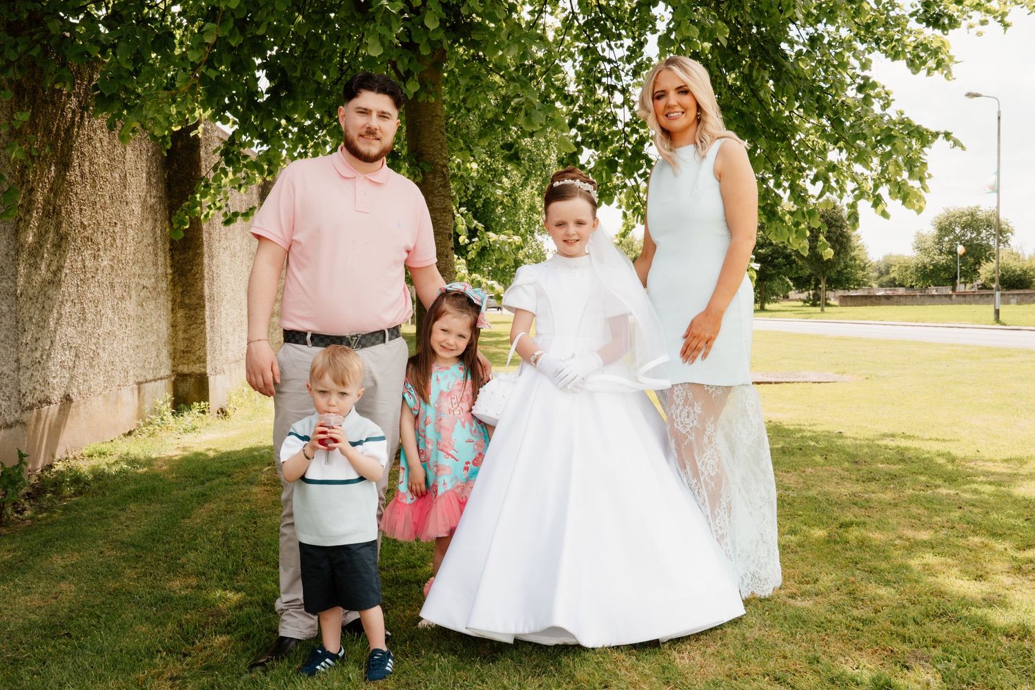 Family of five posing outdoors, girl in white First Communion dress, parents and siblings smiling by a tree.