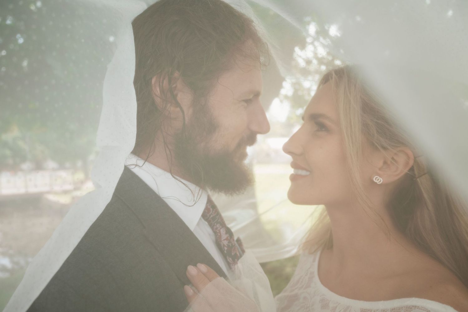 Smiling bride and bearded groom share a tender moment under a soft veil in golden sunlight.