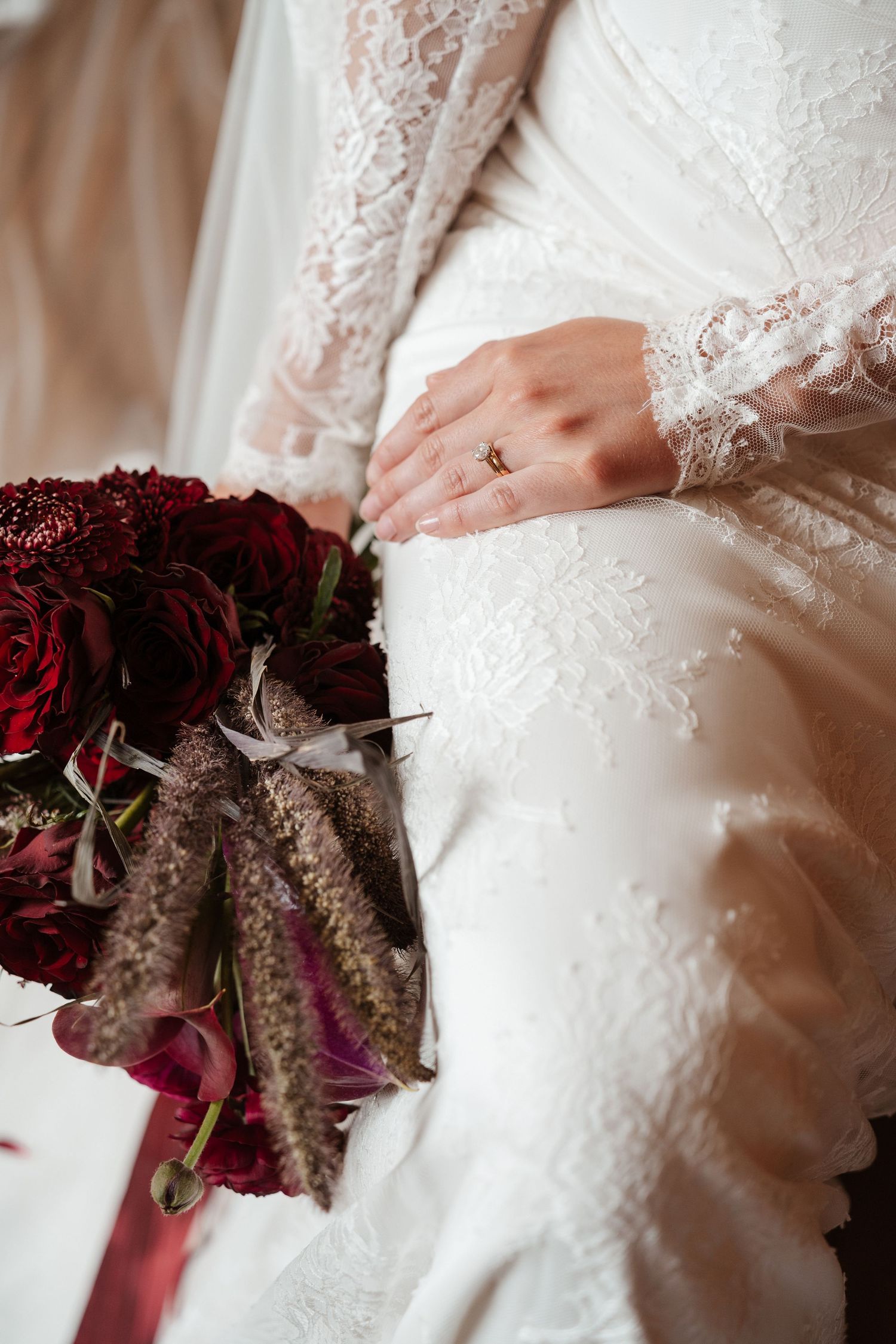 Bride in lace wedding dress holding dark red bouquet with ring on finger.