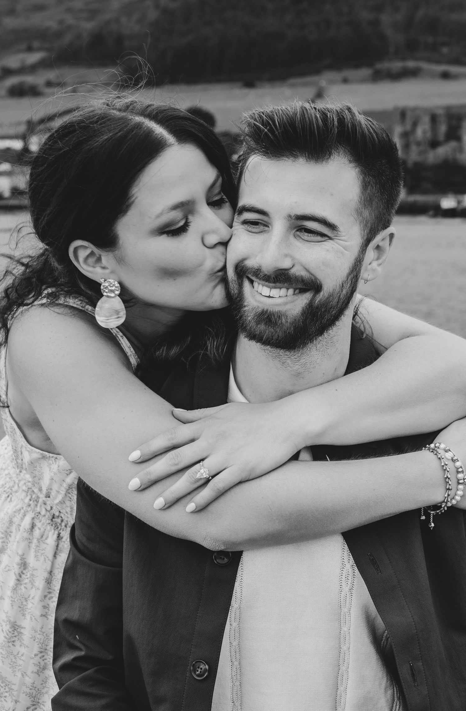 Black and white photo of woman kissing smiling bearded man on cheek while hugging him outdoors.