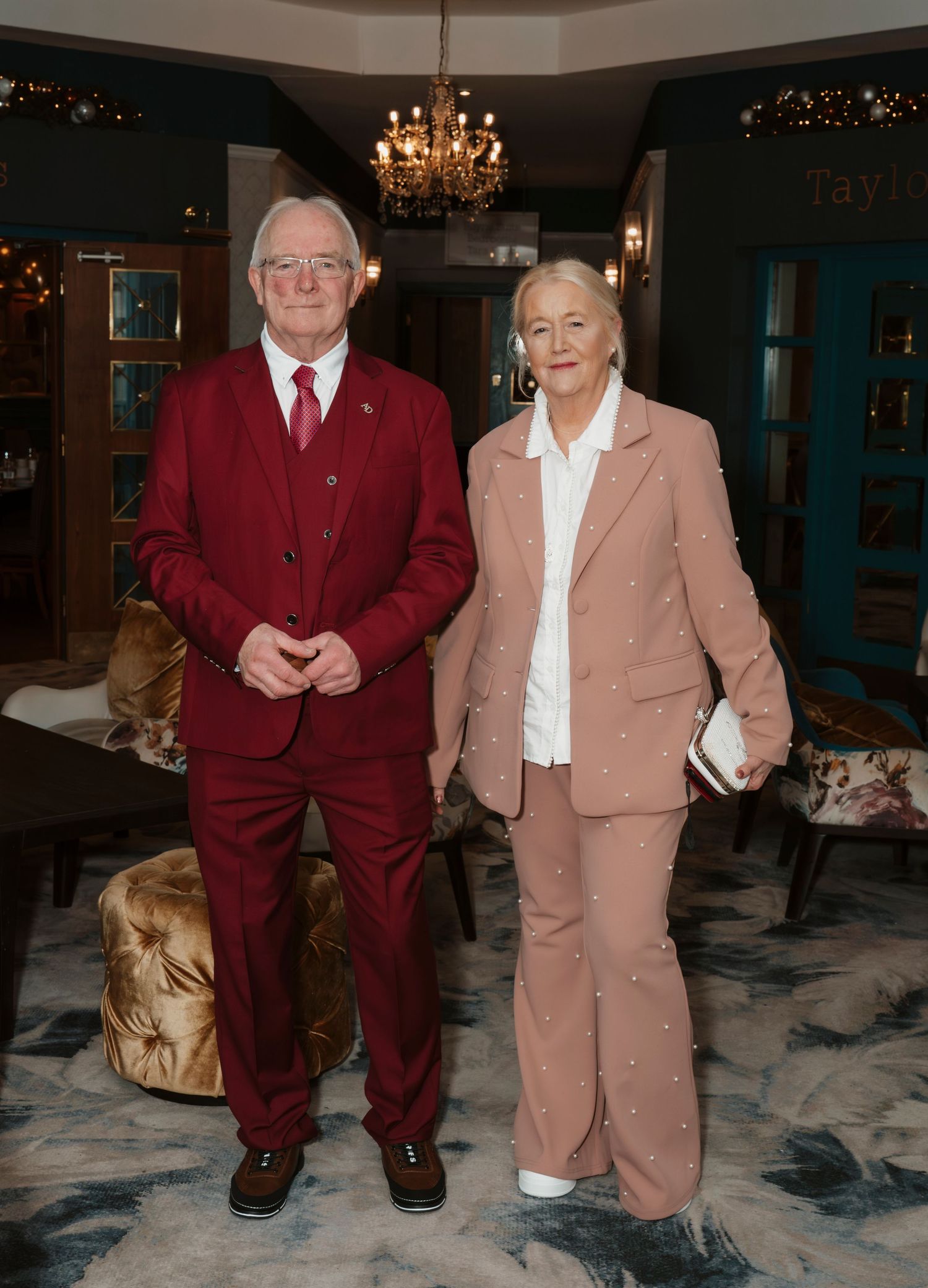 Elegant couple posing in a stylish venue, man in burgundy suit, woman in pink pearl-embellished blazer and trousers.