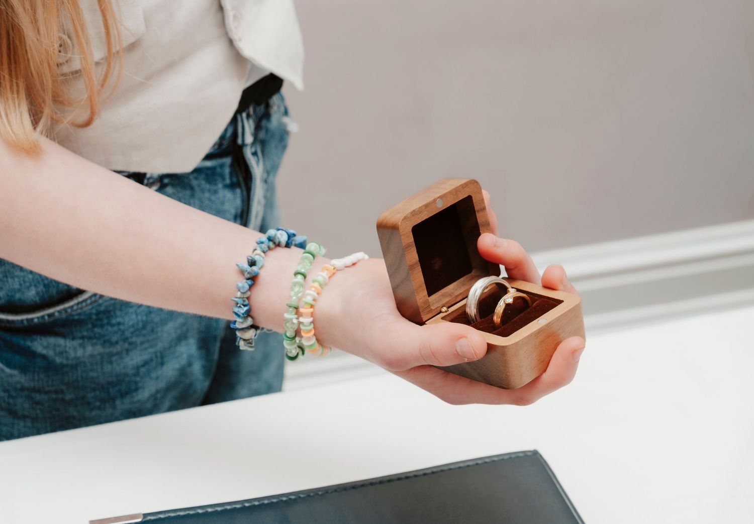 Woman holding a wooden ring box with two rings, wearing colorful beaded bracelets on her wrist.