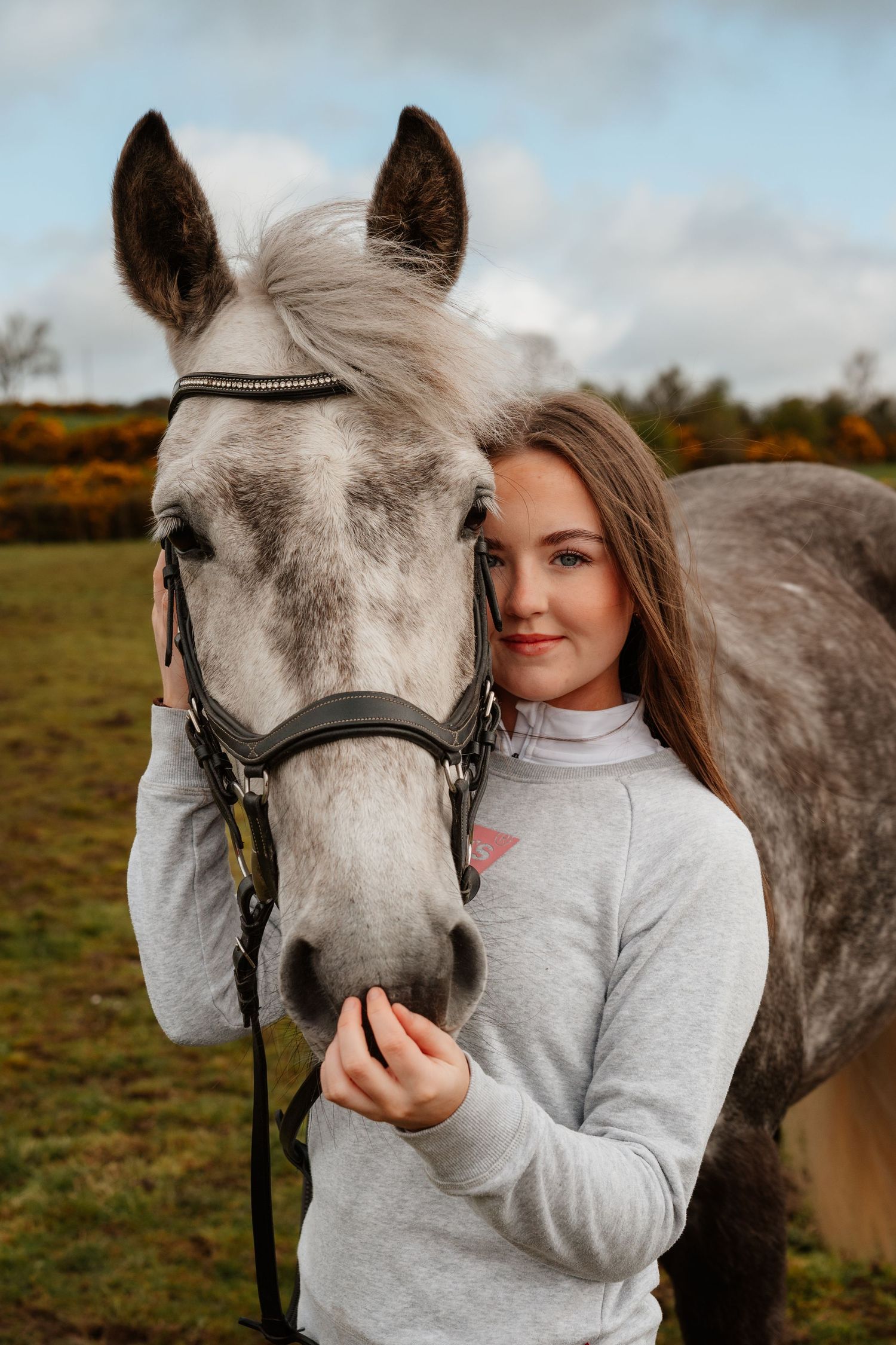 Young woman in gray sweater smiling beside a dapple gray horse wearing a bridle in a green field.