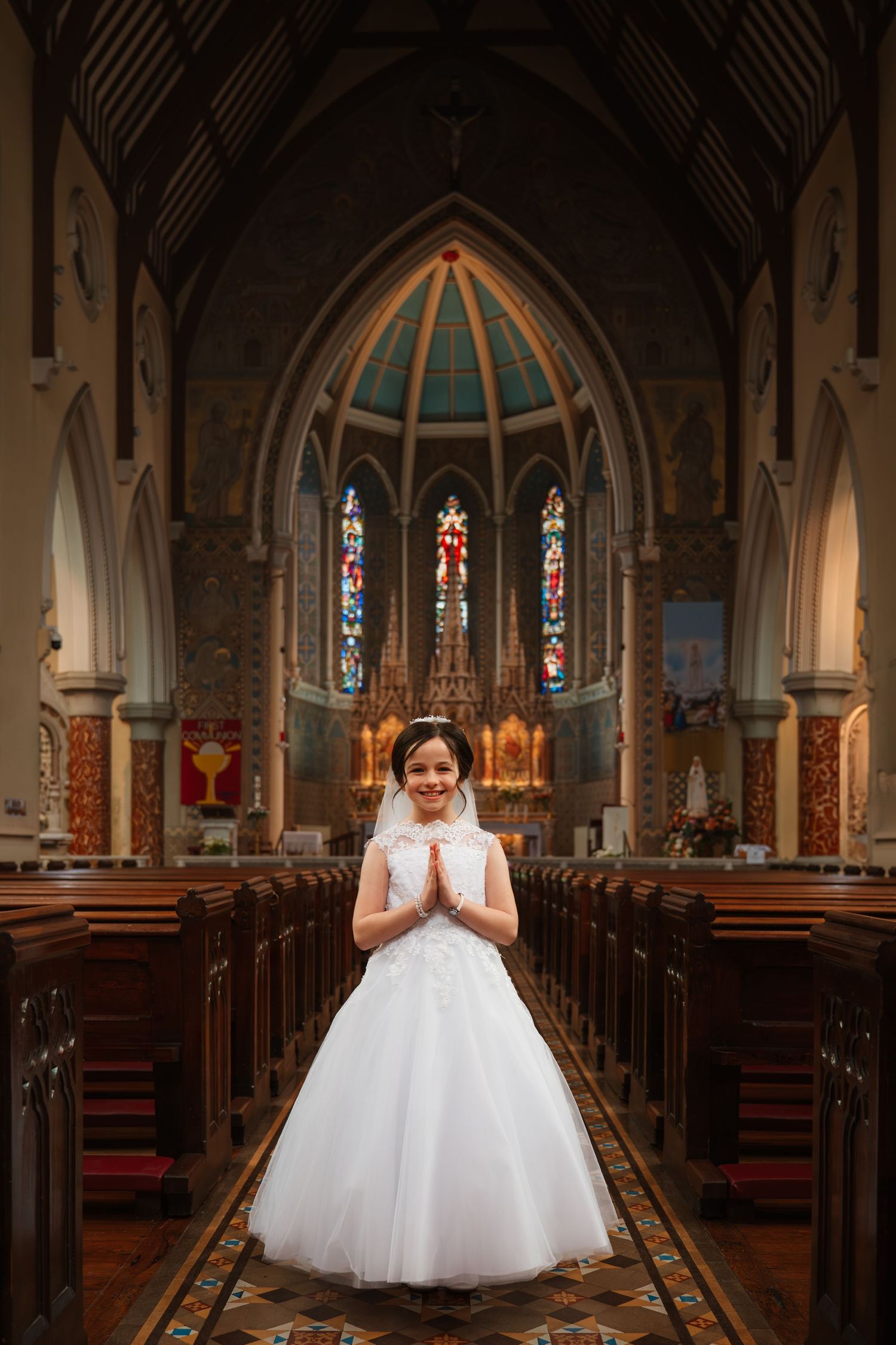 Young girl in white First Communion dress and veil stands with hands clasped in prayer inside a Catholic church.